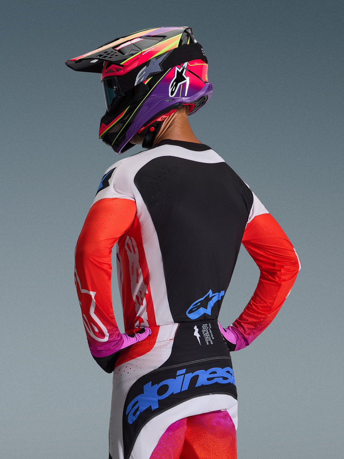 Motocross-Fahrer von hinten im Alpinestars 2026 Techstar Knif Jersey in Light Gray Orange Fluo Black, mit schwarzer Rückenpartie und lasergeschnittener Belüftung, orangefarbenen Ärmeln und hellgrauen Seiteneinsätzen, kombiniert mit einer passenden Hose und einem farbenfrohen Helm.