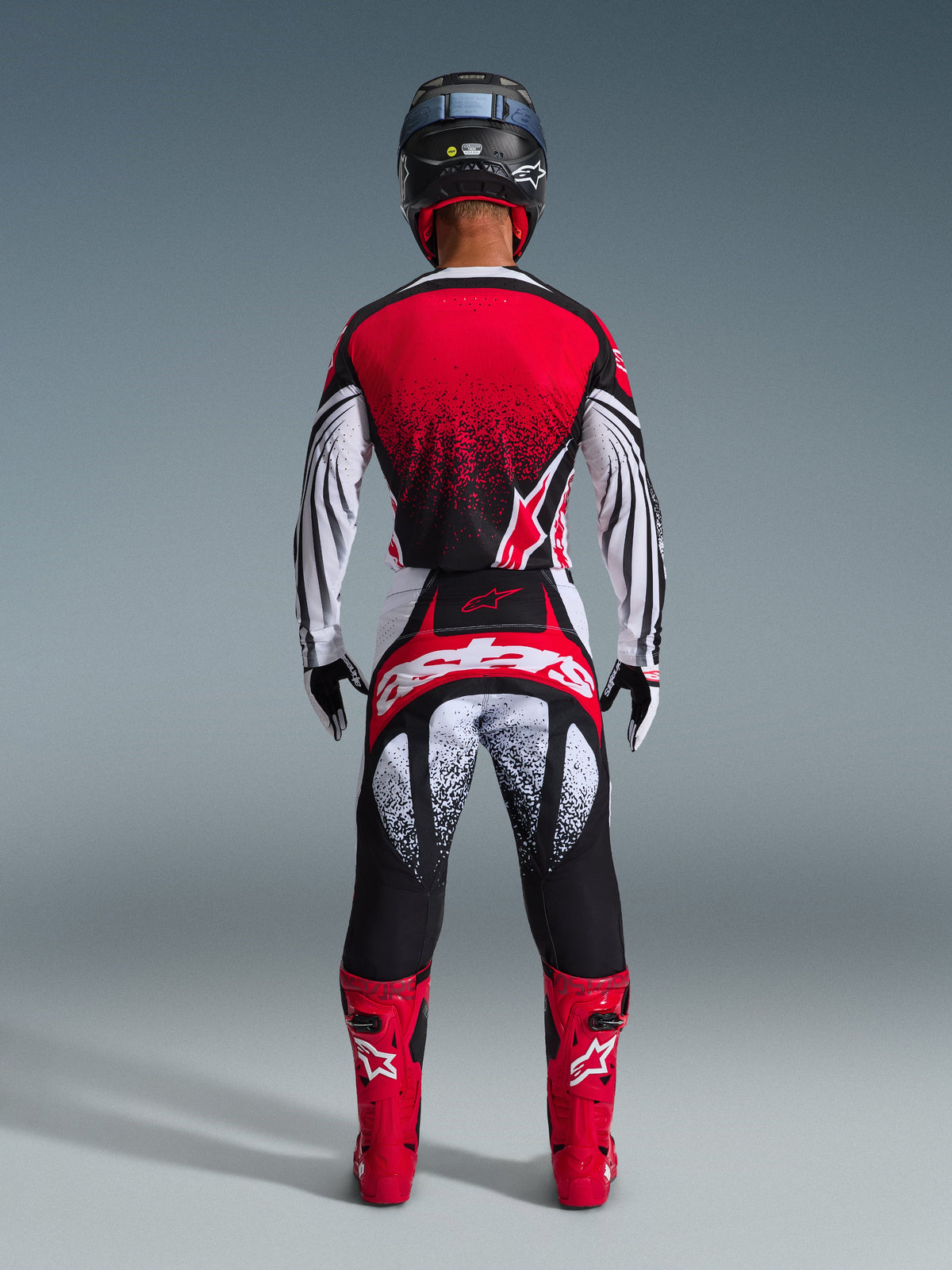 Motocross-Fahrer von hinten im Alpinestars 2026 Techstar Nomur Gear Set, rot-weißes Trikot mit schwarzem Sprenkel-Farbverlauf, passende weiß-rote Hose mit schwarzen Akzenten, leuchtend rote Stiefel und schwarzer Helm, abgestimmte Offroad-Rennausrüstung.