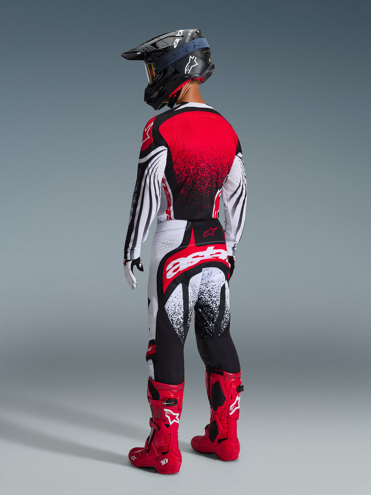 Motocross-Fahrer in der Rückansicht mit dem Alpinestars 2026 Techstar Nomur Gear Set, Trikot in Rot, Weiß und Schwarz mit gepunktetem Farbverlauf, passende Hose mit auffälligem „astars“-Branding, schwarzer Helm und rote Motocross-Stiefel, professionelle Offroad-Rennausrüstung