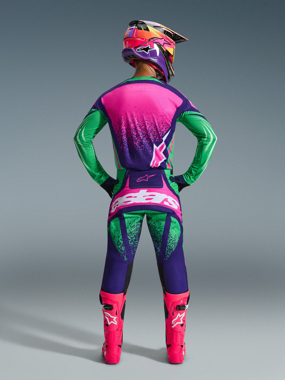 Alpinestars 2026 Techstar Nomur Gear Set, Motocross-Fahrer in der Rückansicht mit Trikot und Hose in dunklem Lila, Grün und Fluo-Pink, dazu passender mehrfarbiger Helm, leuchtend fluo-pinke Stiefel, abgestimmtes professionelles Rennoutfit für Offroad-Performance