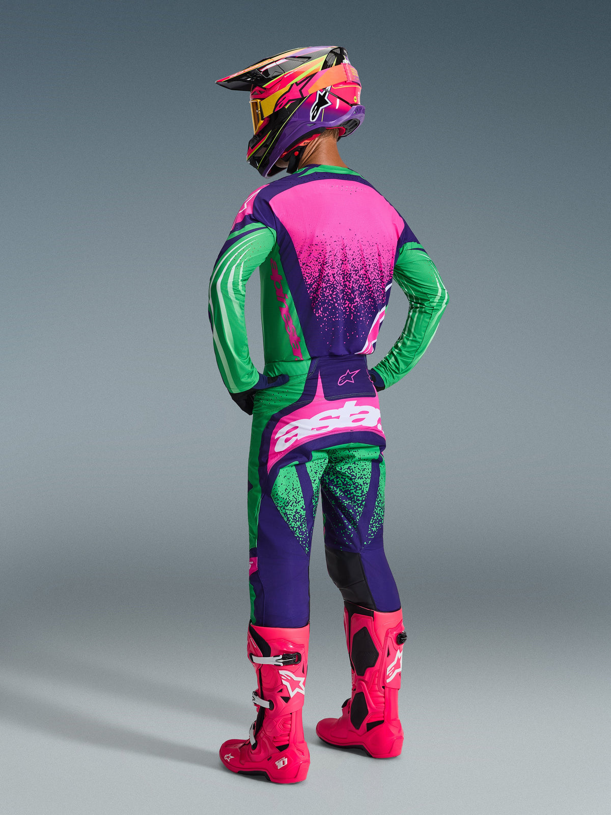 Motocross-Fahrer im 2026 Techstar Nomur Bekleidungsset, Deep Purple Green und Pink Fluo, Trikot mit pinker Rückseite und punktierter Textur, passende Hose mit weißen Logos, leuchtend pinke Fluo-Stiefel und bunter Helm, Ganzkörper-Rückansicht für Offroad-Rennperformance