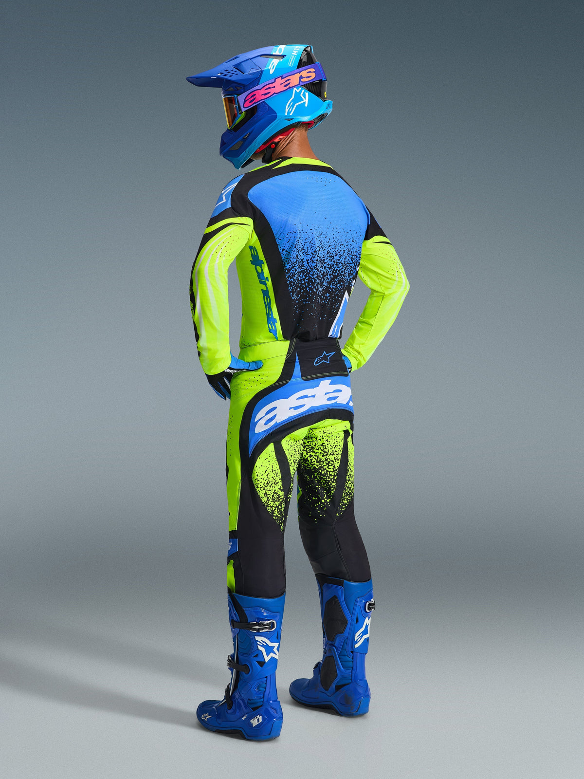 Motocross-Fahrer von hinten in kompletter Rennausrüstung, 2026 Techstar Nomur Trikot in Gelb Fluo und Blau, dazu passende gelb-schwarze Hose mit blauen Akzenten, blauer Helm mit pinkem Alpinestars-Logo und blaue Stiefel, Studioaufnahme, die das Rückendesign und die athletische Passform hervorhebt.
