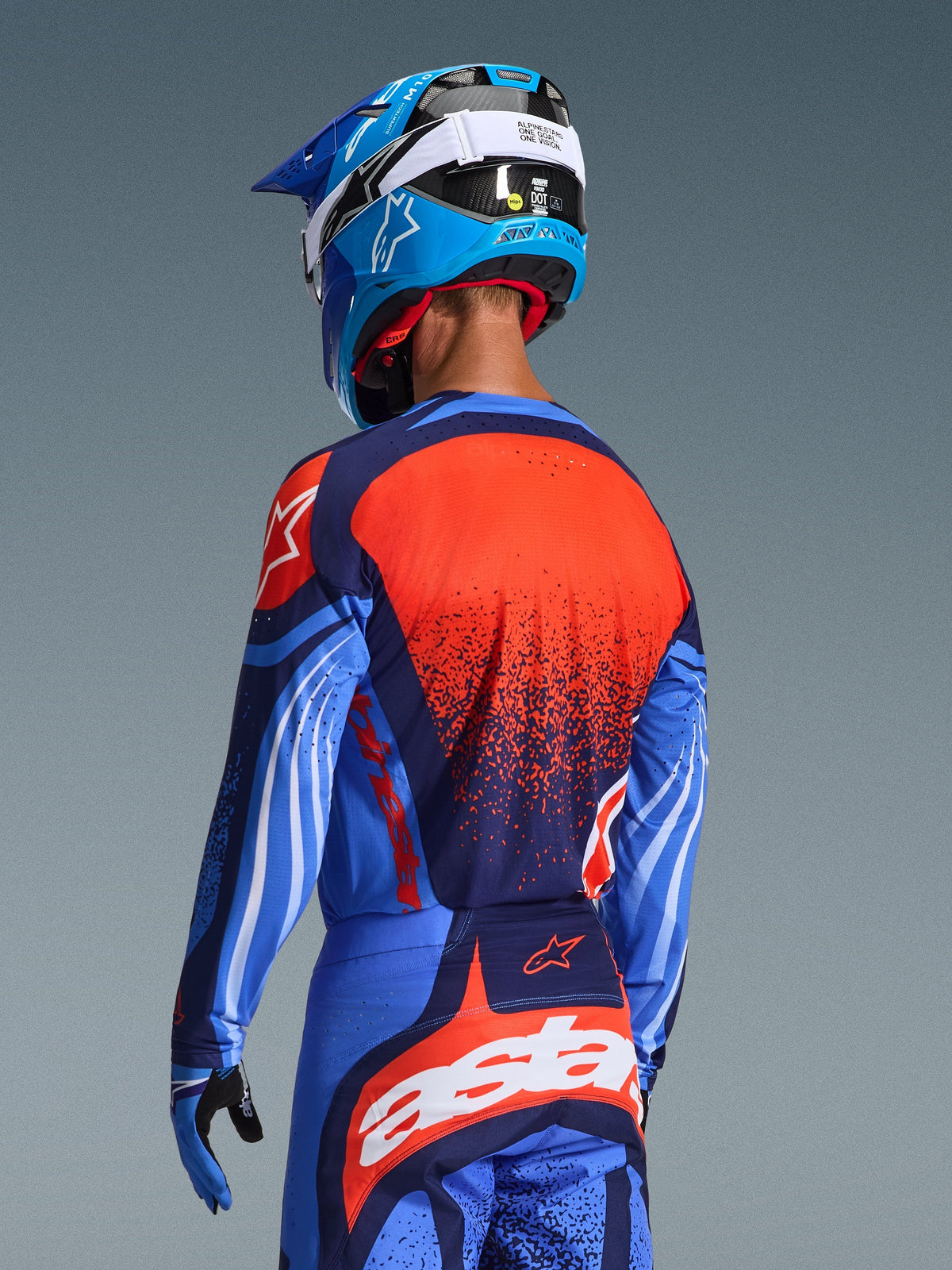 Motocross rider from the back wearing the Alpinestars 2026 Techstar Nomur Jersey and matching pants, Dark Navy and UCLA Blue with vibrant orange accents, featuring a large star graphic and laser-cut ventilation on the jersey, paired with a blue and white helmet for a complete off-road racing kit