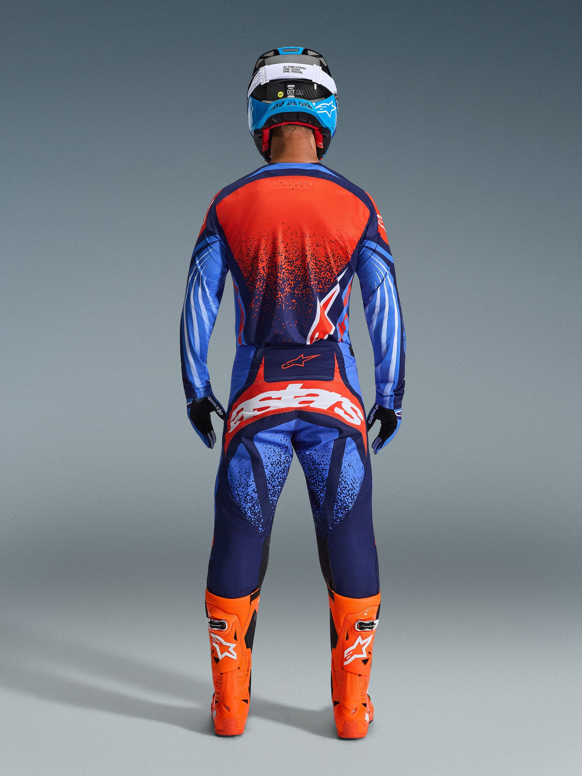 Motocross rider from behind wearing the Alpinestars 2026 Techstar Nomur Gear Set in Dark Navy, Orange, and UCLA Blue, jersey with a speckled orange back, matching pants with bold white branding on the seat, bright orange boots, and a blue and white helmet, coordinated off-road racing kit.