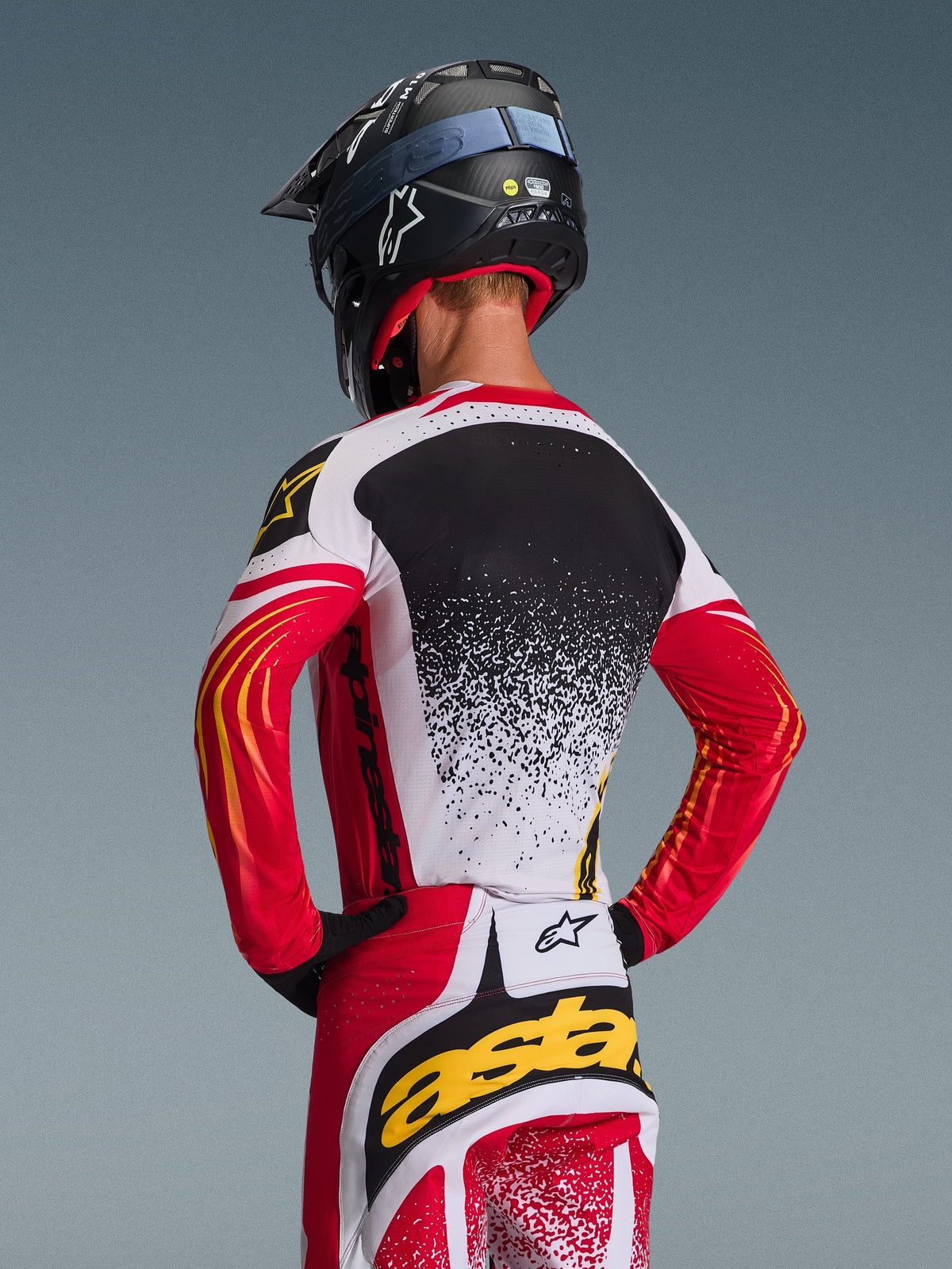 Motocross rider from behind wearing the Alpinestars 2026 Techstar Nomur Jersey and matching pants, Red White and Black with yellow accents, featuring a speckled gradient design on the back, black Alpinestars helmet and black gloves, coordinated professional gear for off-road racing
