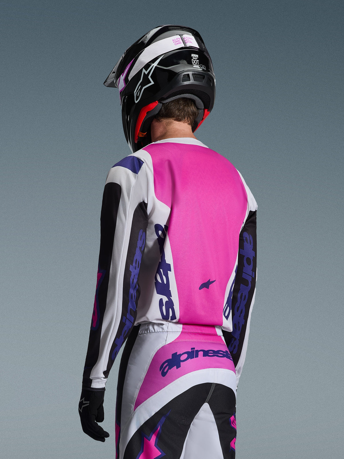 Motocross-Fahrer in der Rückansicht mit dem Alpinestars 2026 Fluid Grid Trikot in Schwarz, Weiß und Lila, passende Hose mit markantem Branding, schwarzer Helm mit pinkfarbenen Akzenten und schwarze Handschuhe, komplett aufeinander abgestimmte Ausrüstung für Offroad-Performance.