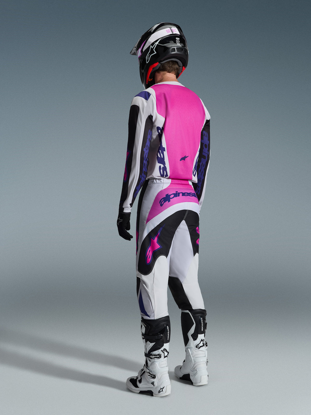 Alpinestars 2026 Fluid Grid Gear Set, Rear view of a motocross rider wearing a pink, white, and black coordinated outfit, jersey with large pink back panel and purple branding, matching pants with black and pink accents, black helmet, and white and black off-road boots.