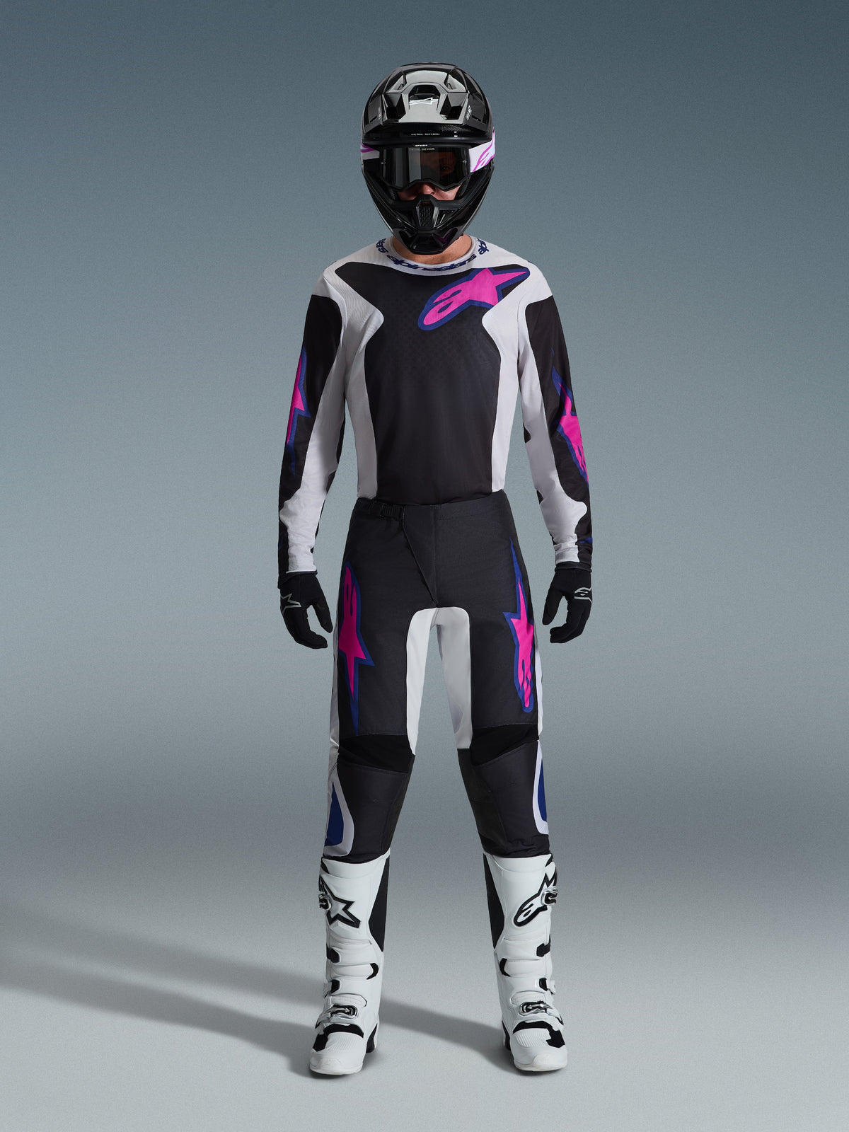 Motocross-Fahrer in kompletter Rennausrüstung, Alpinestars 2026 Fluid Grid Trikot und passende Hose in Schwarz und Hellgrau mit leuchtend lila Akzenten, schwarzer Helm mit lila Goggles, weiße Motocross-Stiefel und schwarze Handschuhe, abgestimmtes Outfit für Offroad-Performance