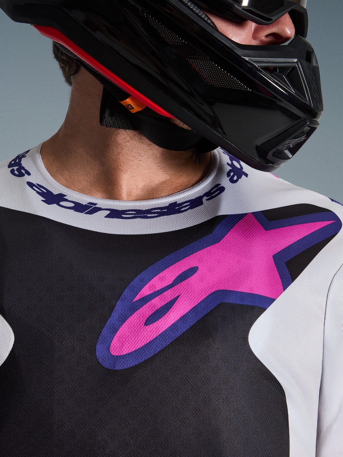 Alpinestars 2026 Fluid Grid Trikot, Motocross-Trikot, Schwarz, Hellgrau und Lila, Nahaufnahme des Brust- und Halsbereichs mit großem pinkfarbenem Stern-Logo, Sportkragen mit Markenschriftzug, getragen mit schwarzem Helm, Hochleistungs-Offroad-Ausrüstung