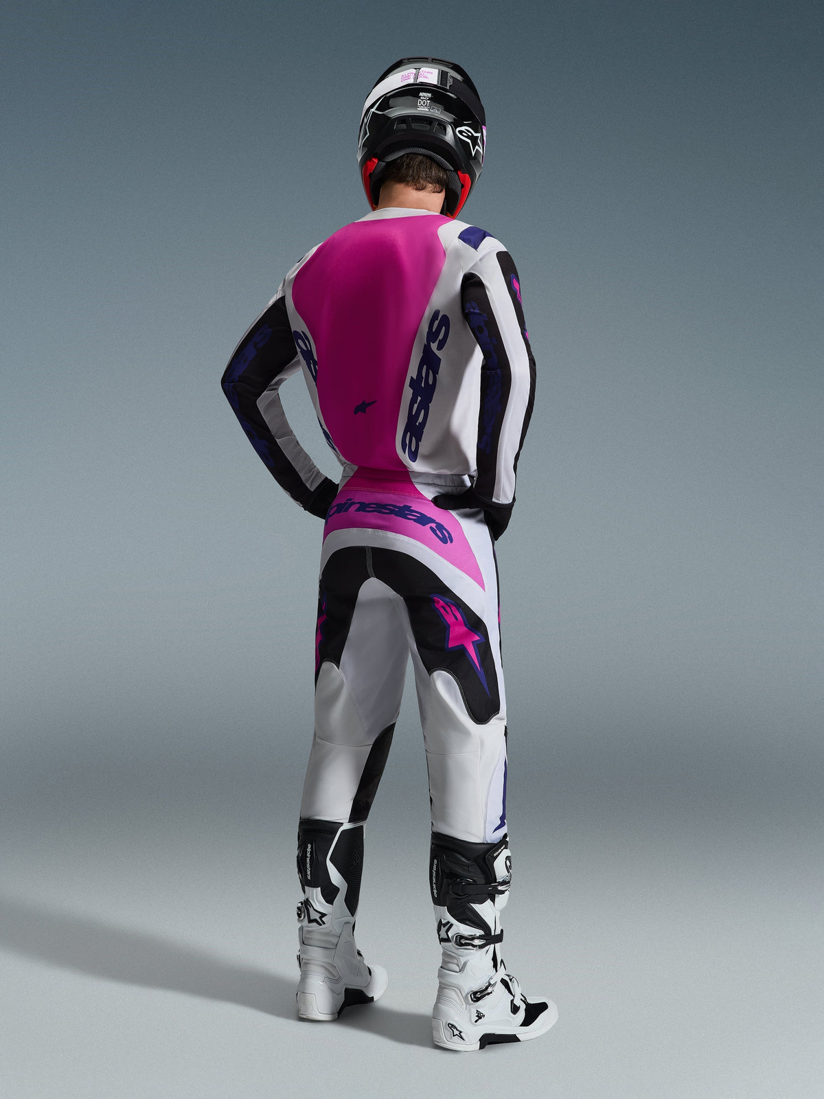 Motocross-Fahrer in der Rückansicht, Alpinestars 2026 Fluid Grid Trikot, weißes Trikot mit großem leuchtend pinkfarbenem Rückenteil und lila Branding, passende weiß-schwarze Hose mit pinkfarbenen Akzenten, weiße Stiefel und schwarzer Helm, abgestimmte Offroad-Rennausrüstung