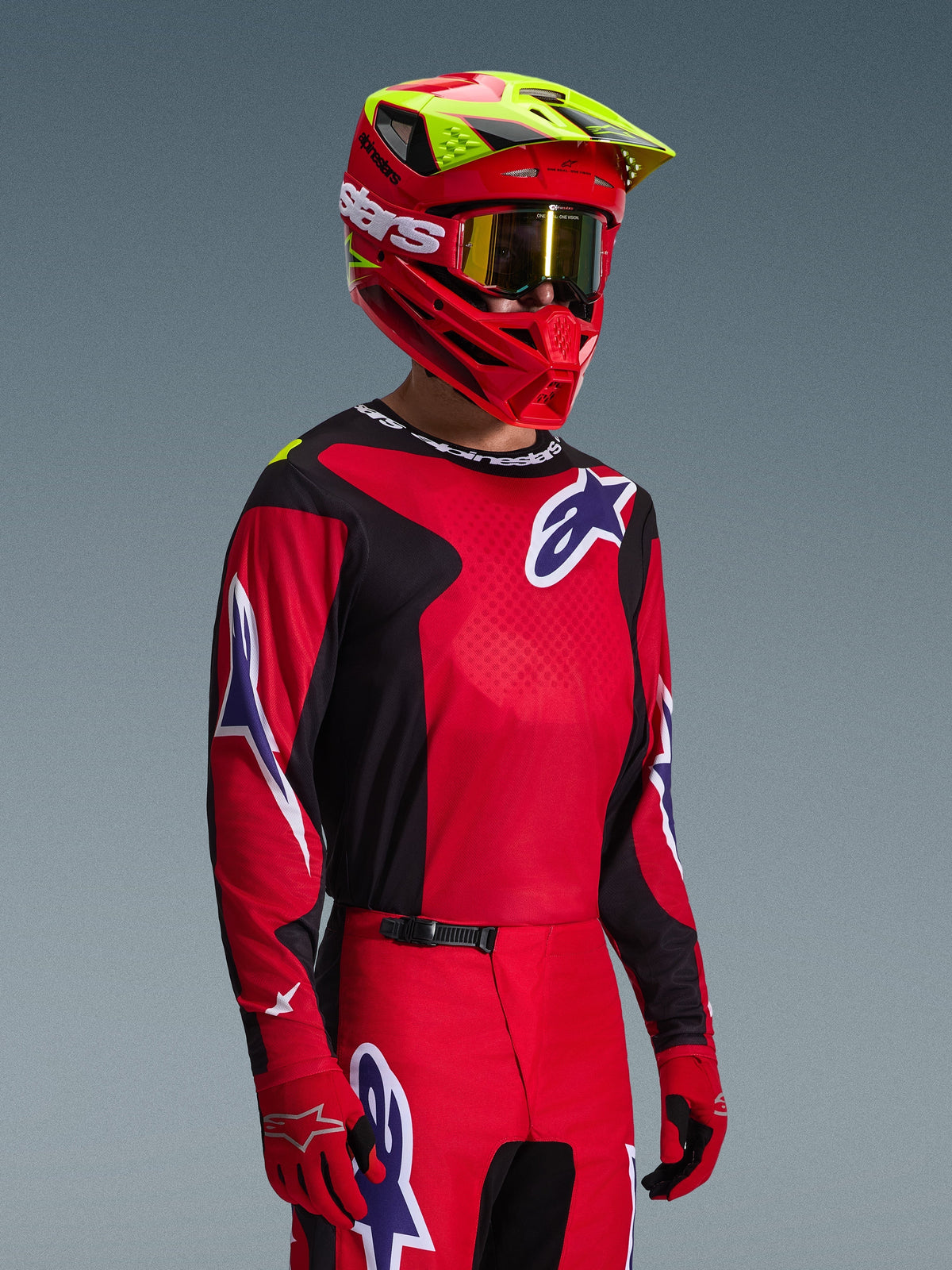 Motocross-Fahrer im Alpinestars 2026 Fluid Grid Bekleidungsset, rotes und schwarzes Jersey mit weißen und blauen Logo-Akzenten, passende rote und schwarze Hose, roter Helm mit gelben Highlights und goldenen Goggles, abgestimmte Offroad-Rennausrüstung