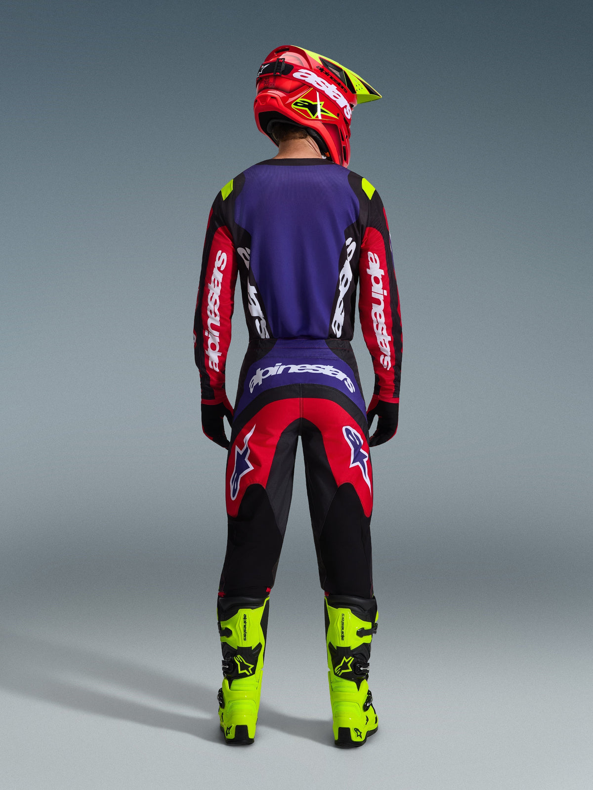 Motocross-Fahrer von hinten im Alpinestars 2026 Fluid Grid Gear Set in Rot, Schwarz und Lila, mit einem lila Rückenteil und roten Ärmeln am Trikot, passender rot-schwarzer Hose, leuchtend neongelben Motocross-Stiefeln und einem rot-neongelben Helm.