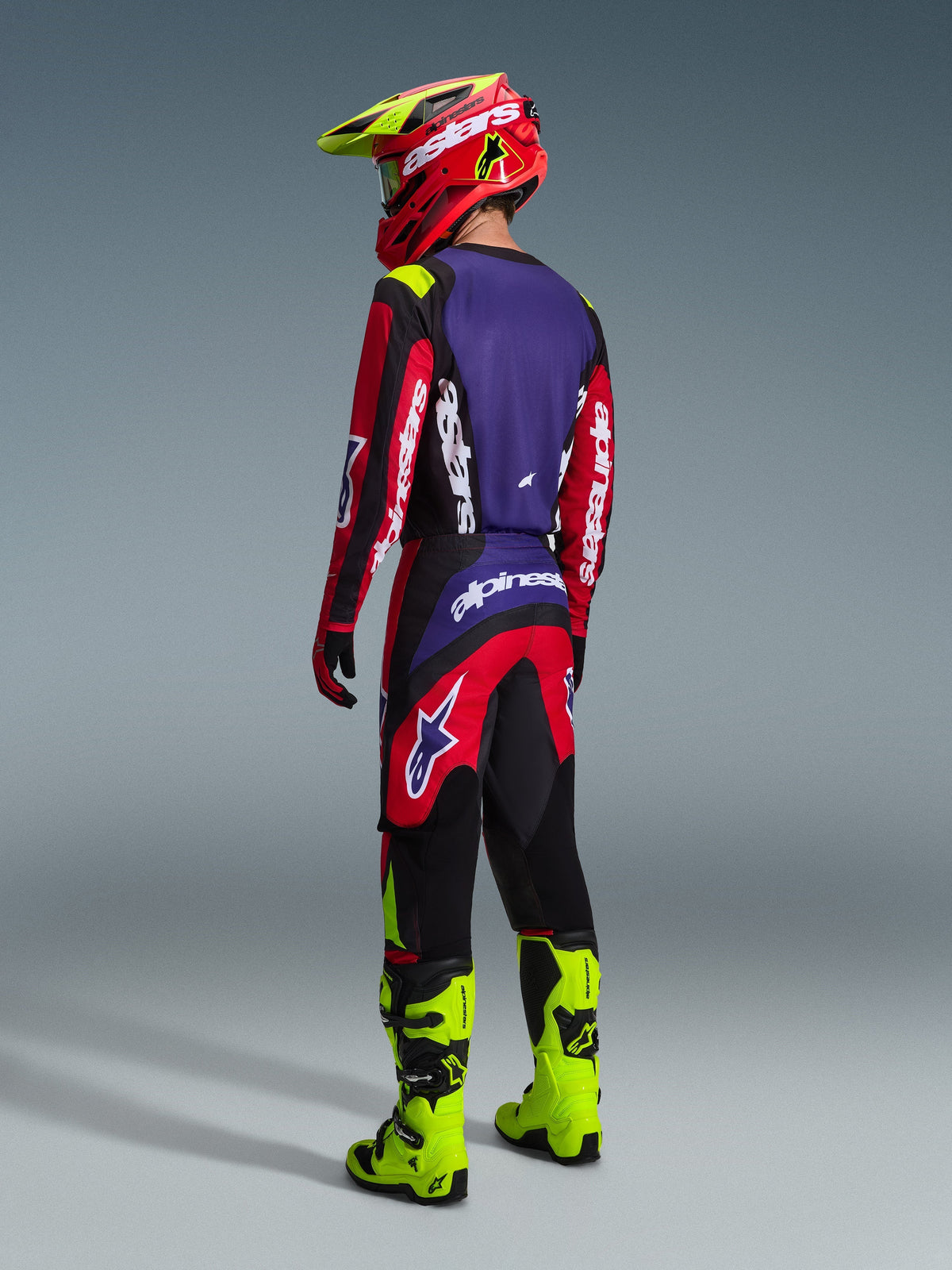 Motocross-Fahrer von hinten im Alpinestars 2026 Fluid Grid Gear Set, Trikot in Rot, Schwarz und Lila mit weißem Branding, rote und schwarze Hose mit lila Akzenten, kombiniert mit einem rot-neongelben Helm und leuchtend neongelben Stiefeln, komplette Offroad-Rennausrüstung.