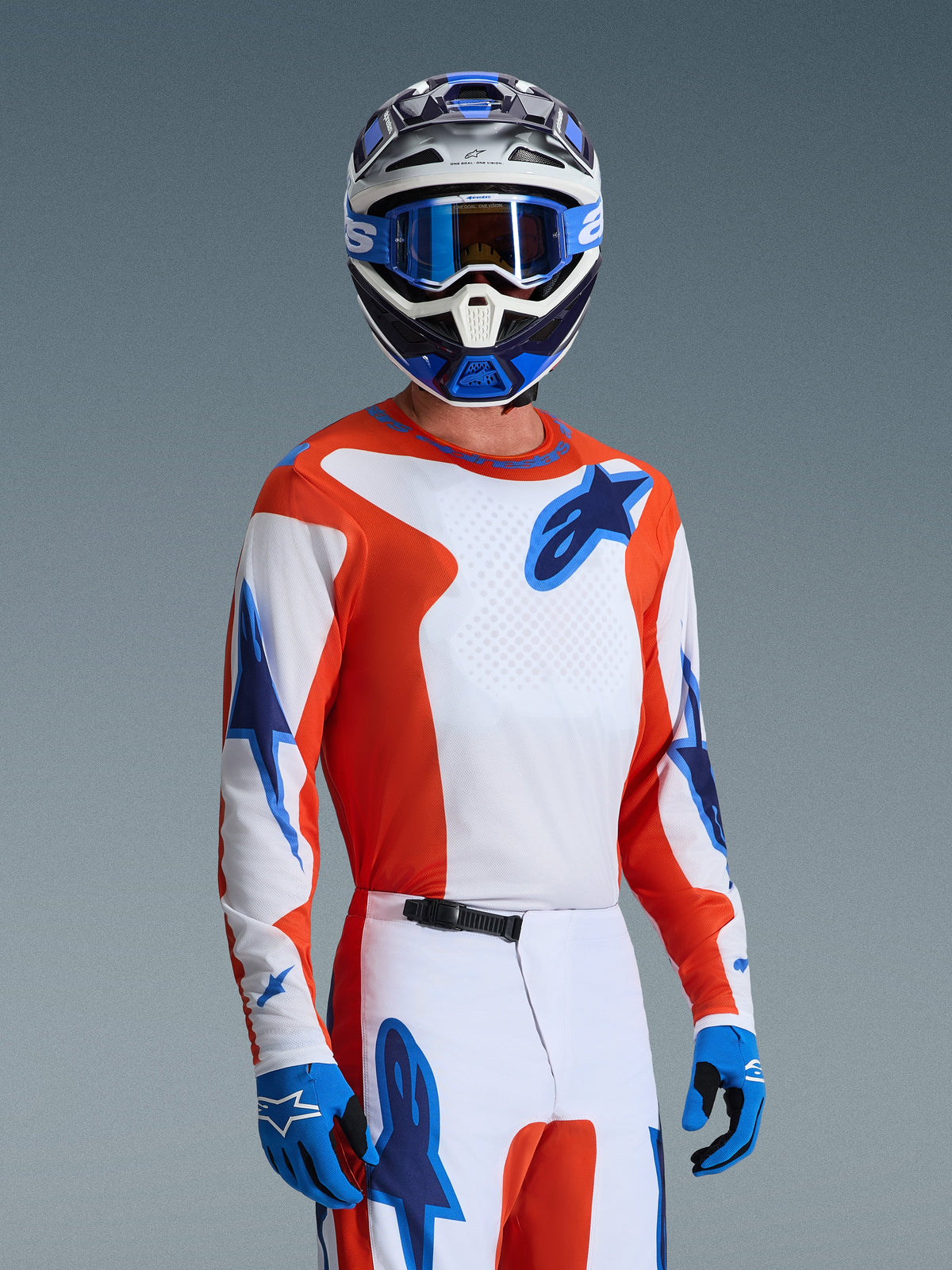 Alpinestars 2026 Fluid Grid Trikot, Motocross-Trikot in Orange UCLA Blue, getragen von einem Fahrer mit passender weiß-oranger Hose, blau-weißem Helm, blauer Brille und blauen Handschuhen, komplettes Offroad-Rennausrüstungs-Set