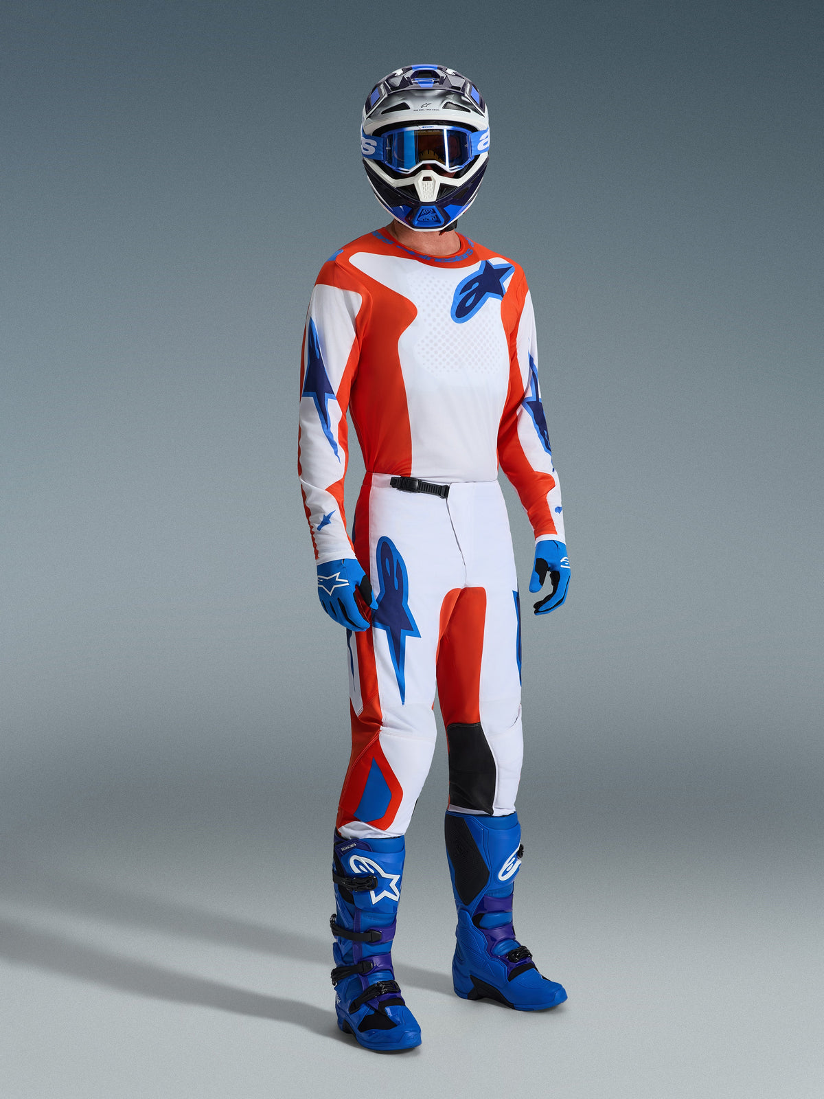 Motocross-Fahrer in kompletter Rennausrüstung, 2026 Fluid Grid Trikot in Orange und UCLA-Blau, passende Hose mit weißen Einsätzen und blauen Logos, blaue Motocross-Stiefel und ein farblich abgestimmter Helm, professionelle Studioaufnahme für den Offroad-Rennsport