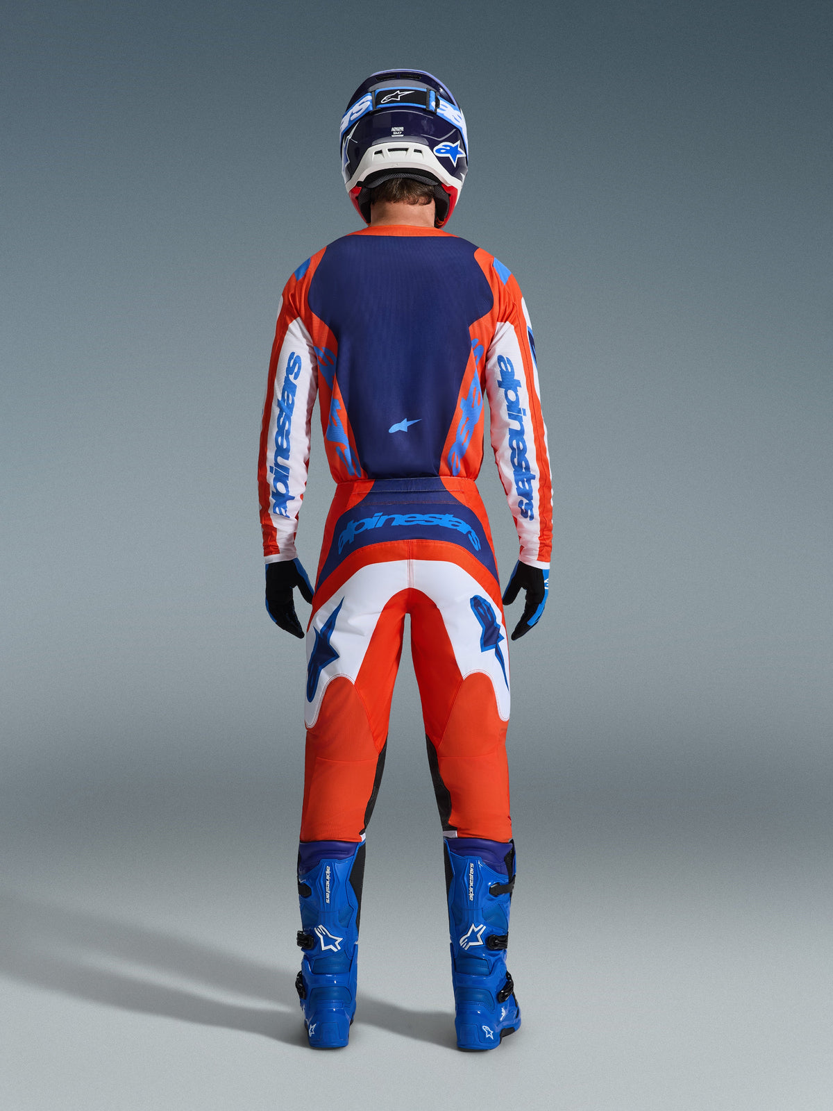 Alpinestars 2026 Fluid Grid Trikot, Motocross-Trikot, Orange und UCLA-Blau, Rückansicht eines Fahrers in kompletter Rennausrüstung, mit dunkelblauem Rückeneinsatz und weißen Ärmeln mit blauem Branding, kombiniert mit passender Hose und blauen Stiefeln.