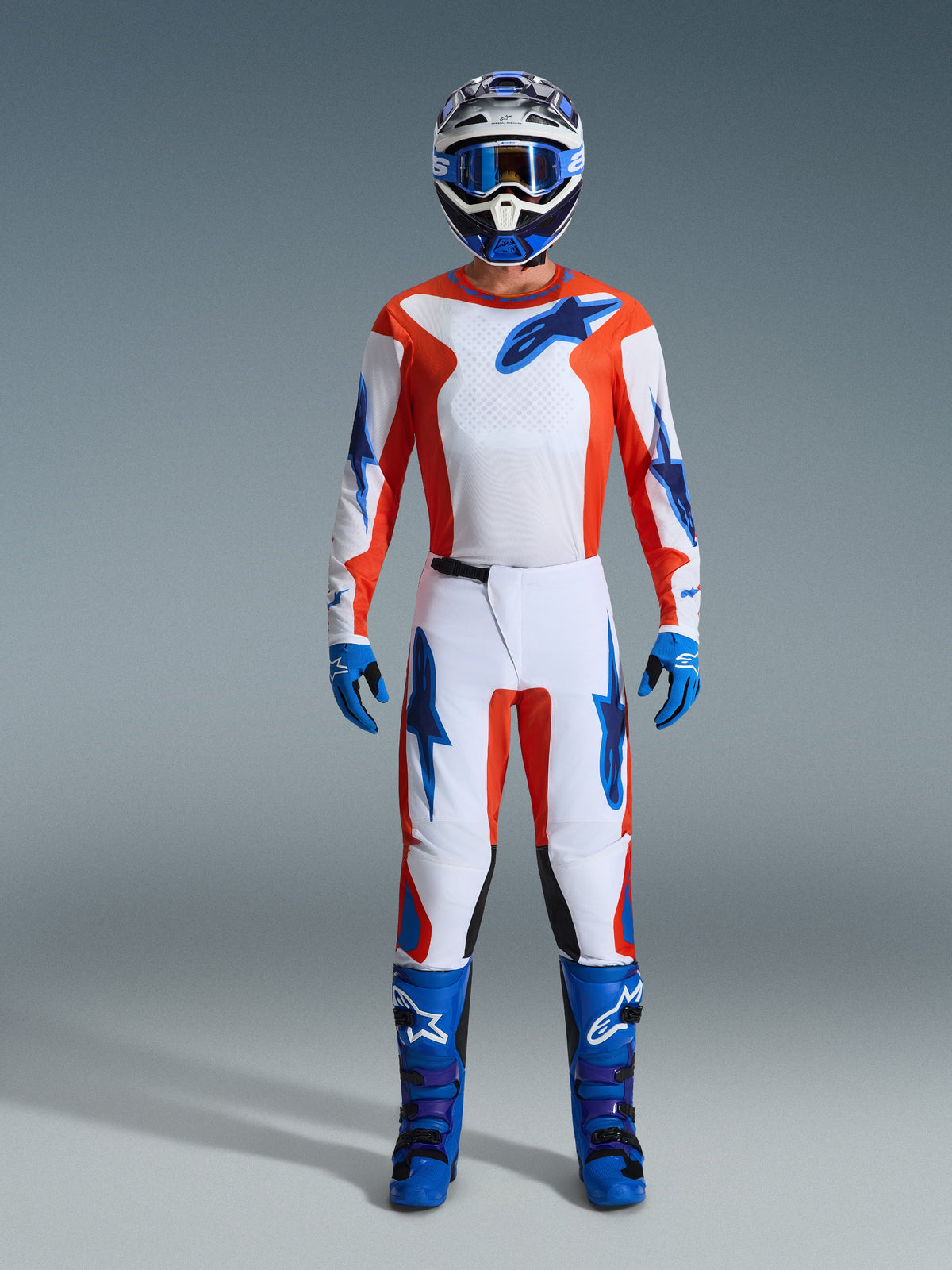 Motocross-Fahrer in kompletter Rennausrüstung, 2026 Fluid Grid Trikot in Orange und UCLA-Blau, passende weiße Hose mit orangefarbenen und blauen Akzenten, blau-weißer Helm mit blauer Schutzbrille, blau-violette Offroad-Stiefel, blaue Handschuhe, abgestimmtes Outfit für Motocross-Rennen