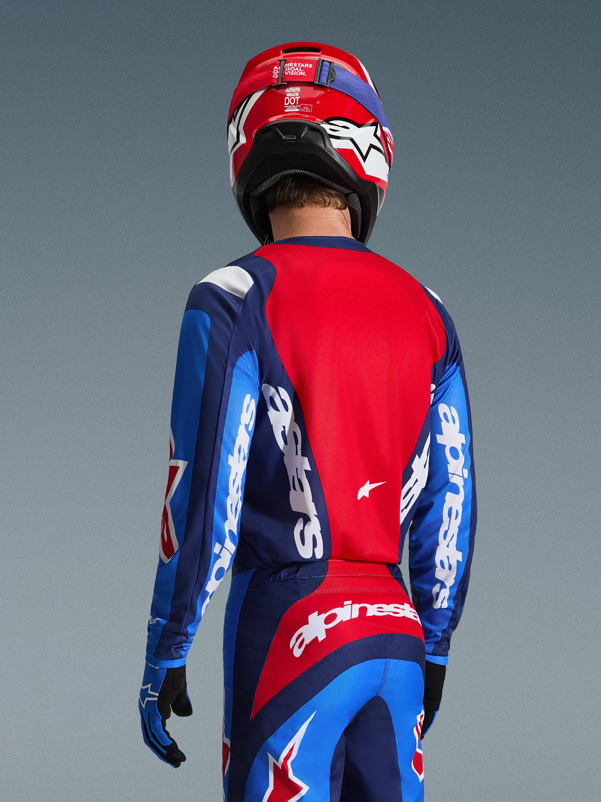 Rückansicht eines Motocross-Fahrers im Alpinestars 2026 Fluid Grid Trikot in Blau und Rot, mit markantem weißem Branding auf den Ärmeln und dem Rücken, kombiniert mit der passenden Motocross-Hose und einem roten Helm für einen kompletten Offroad-Racing-Look