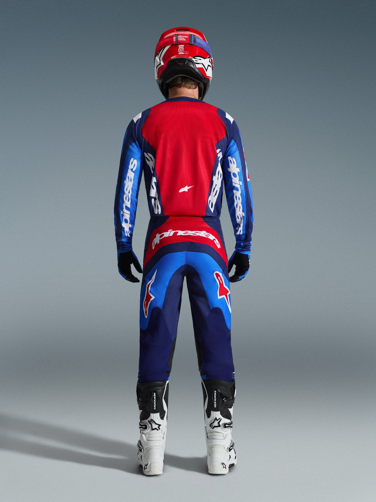 Rückansicht eines Motocross-Fahrers im 2026 Fluid Grid Jersey in Blau und Rot, mit einem großen roten Rückenteil und blauen Ärmeln mit weißem Alpinestars-Schriftzug, kombiniert mit der passenden Hose, weiß-schwarzen Stiefeln und einem roten Helm für ein komplettes Offroad-Rennoutfit.