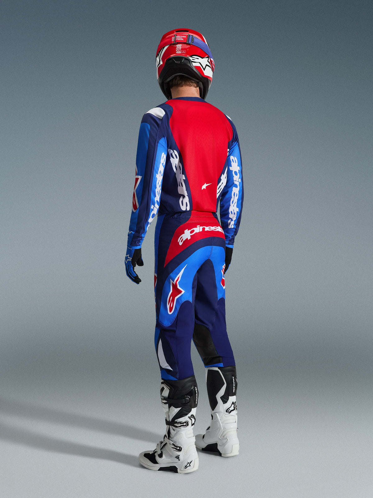 Motocross-Fahrer in der Rückansicht mit dem Alpinestars 2026 Fluid Grid Trikot in Blau und Rot, passender Motocross-Hose mit auffälligem weißen Branding, rotem Helm und weiß-schwarzen Stiefeln, komplette Offroad-Rennausrüstung