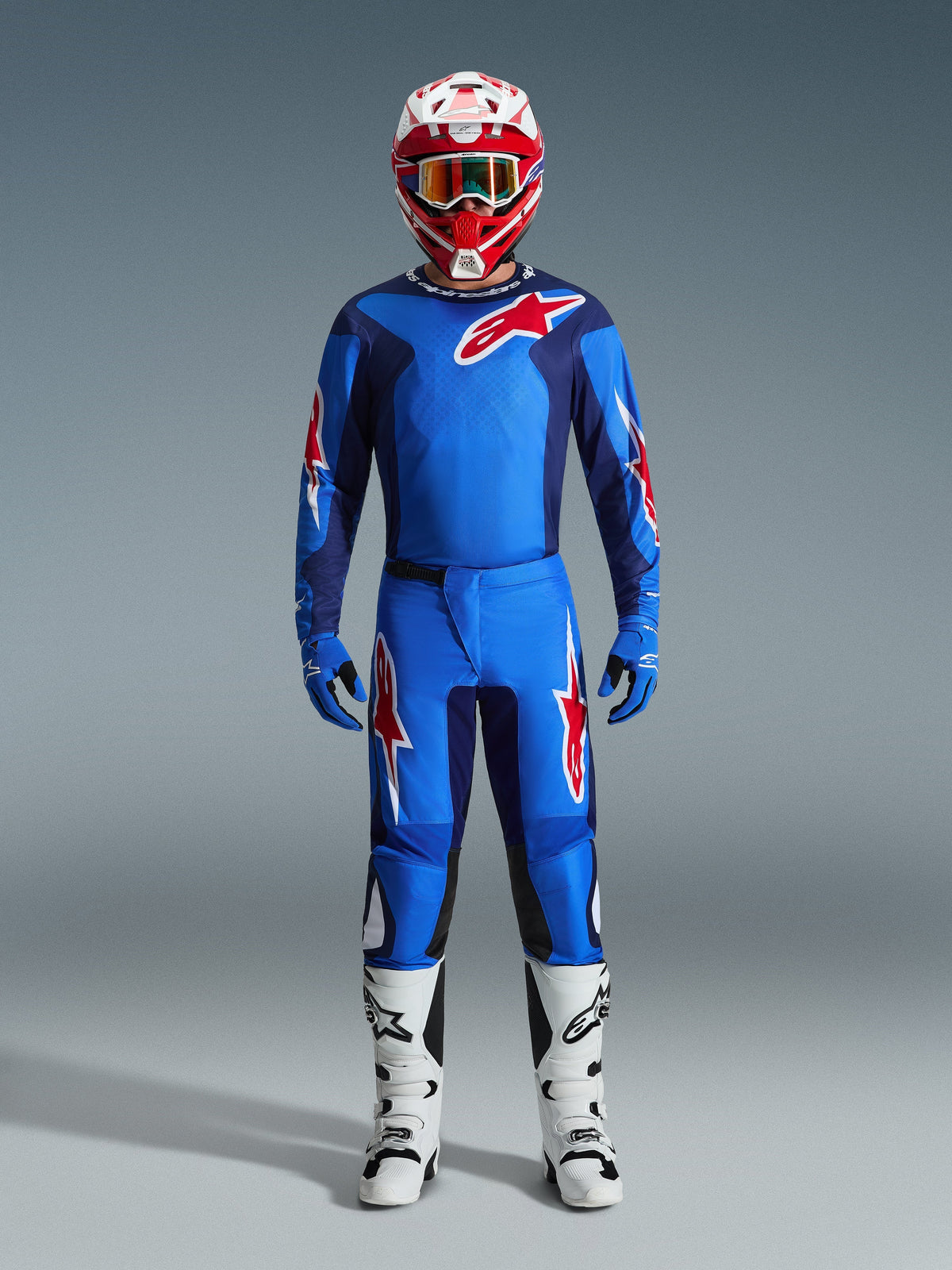 Motocross-Fahrer im Alpinestars 2026 Fluid Grid Trikot in Blau und Rot, passende blaue Hose mit weiß-roten Stern-Logos, rot-weißer Helm mit verspiegelter Schutzbrille, blaue Handschuhe und weiß-schwarze Rennstiefel, komplette Offroad-Ausrüstung.