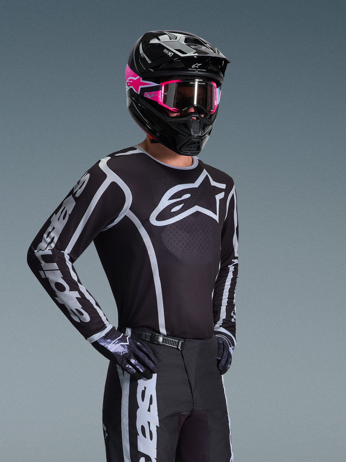 Motocross-Fahrer im Alpinestars 2026 Fluid Apex Trikot, Schwarz und Grau mit großem weißen Logo auf der Brust und geometrischen Grafiken auf den Ärmeln, kombiniert mit einer schwarzen Hose, schwarzen Handschuhen sowie einem schwarzen Helm mit pinkfarbenen Akzenten und pinker Brille