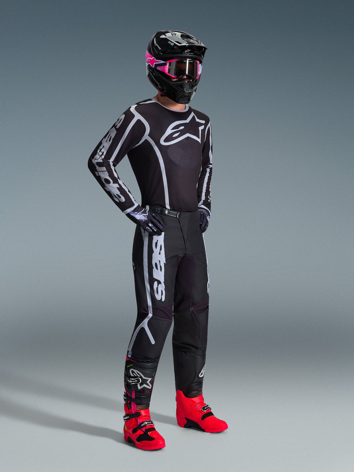 Alpinestars 2026 Fluid Apex Bekleidungsset, Motocross-Fahrer in schwarzem Jersey und Hose mit weiß-grauen Grafikakzenten, schwarzer Helm mit pinker Brille und leuchtend pinken Rennstiefeln, koordiniertes professionelles Motocross-Outfit