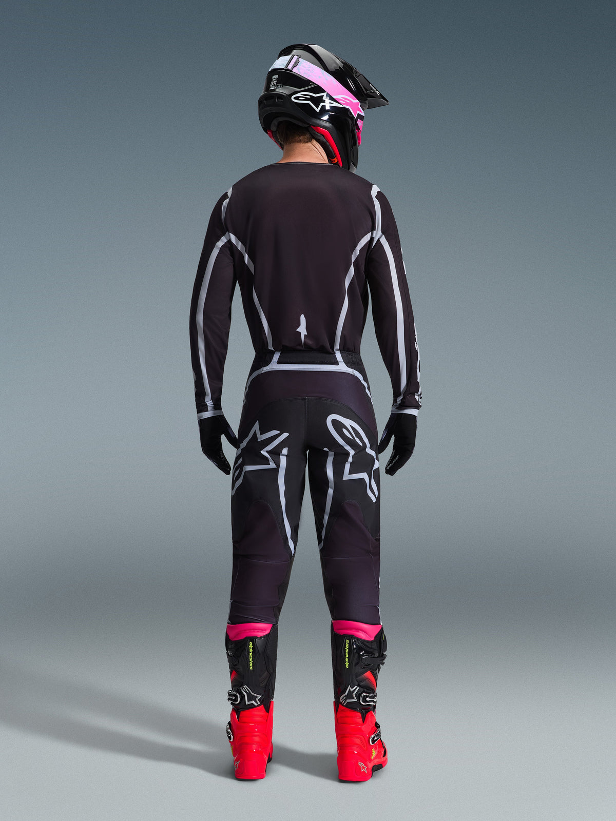 Alpinestars 2026 Fluid Apex Gear Set, Motocross-Fahrer von hinten gesehen, schwarzes Jersey und Hose mit grauen geometrischen Linien und Stern-Logos, schwarzer Helm mit pinkfarbenen Akzenten, leuchtend rote und schwarze Stiefel, abgestimmte Motocross-Rennausrüstung