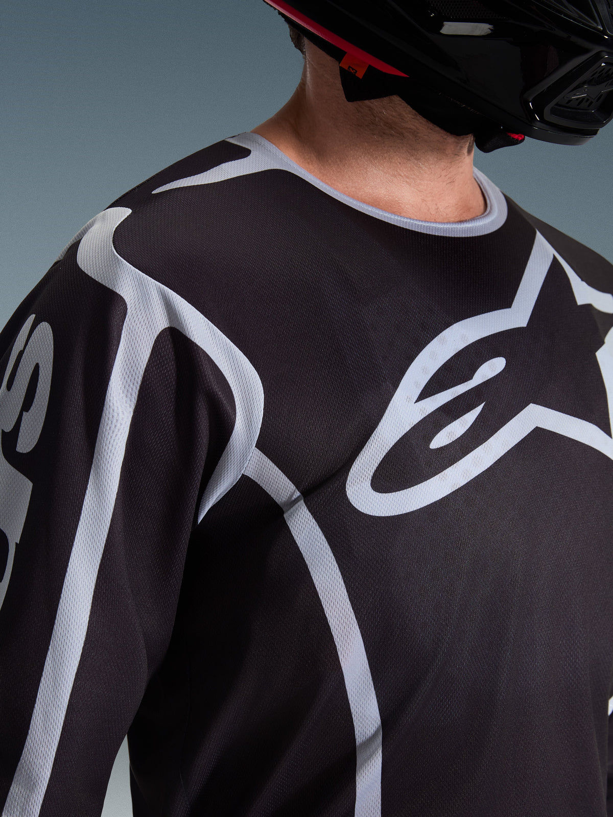 Alpinestars 2026 Fluid Apex Jersey, Motocross-Jersey, Schwarz und Grau, mit großem Alpinestars-Logo auf der Brust, Sportkragen, getragen mit einem schwarzen Helm, Performance-Ausrüstung für den Offroad-Sport