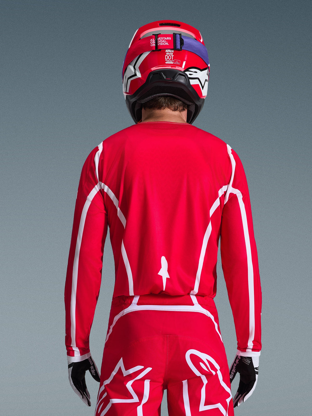 Rear view of a motocross rider wearing the Alpinestars 2026 Fluid Apex Jersey in Red and White, featuring geometric white patterns, styled with matching motocross pants and a red helmet, complete racing gear for off-road performance.