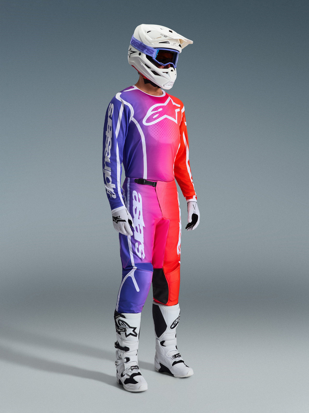 Alpinestars 2026 Fluid Apex Gear Set, Motocross-Fahrer in einem kompletten Rennoutfit mit einem leuchtenden Farbverlauf in Lila, Pink und Rot, weißer Helm mit lila Brille, weiße und schwarze Stiefel, weiße Handschuhe, koordinierter sportlicher Look für Offroad-Performance