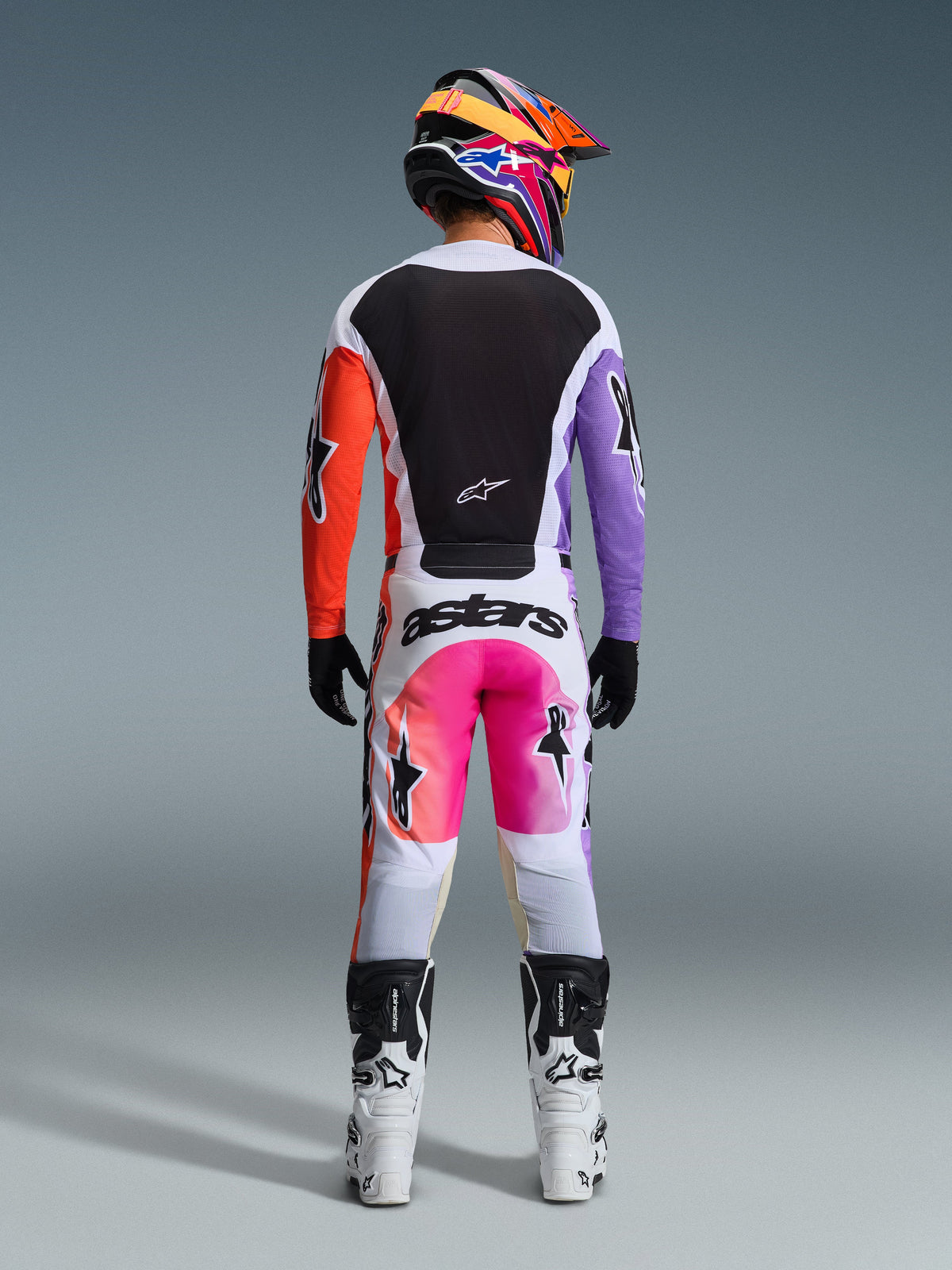 Alpinestars 2026 Racer Air Portl Gear Set, Rückansicht eines Motocross-Fahrers, Jersey mit schwarzem Rücken sowie orangefarbenen und lila Ärmeln, weiße Hose mit pink-orangefarbenem Farbverlauf und markantem „astars“-Branding, farblich abgestimmter mehrfarbiger Helm und weiße Stiefel, professionelle Offroad-Rennausrüstung
