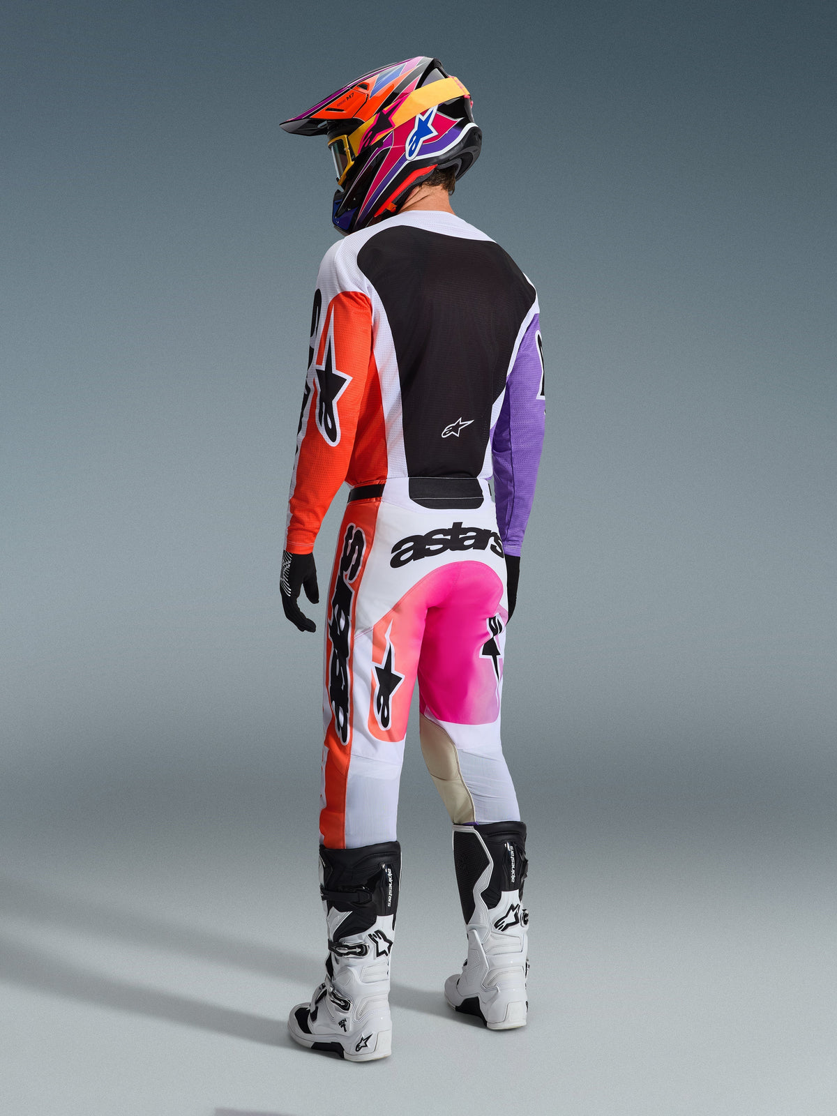 Motocross-Fahrer von hinten in der Alpinestars 2026 Racer Air Portl Hose in Multicolor, mit weißen, orangefarbenen und lila Paneelen sowie neonpinken Akzenten und markantem schwarzen Branding, kombiniert mit passendem Trikot, bunt gemustertem Helm und weiß-schwarzen Stiefeln.