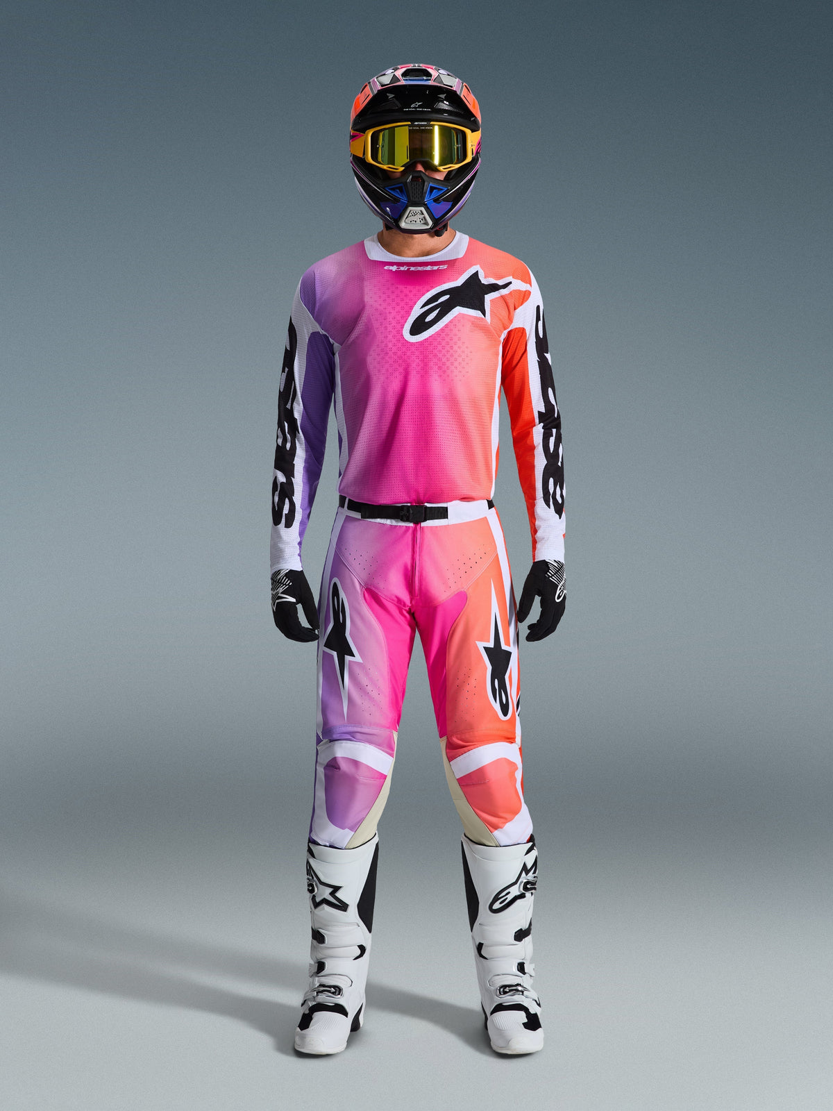 Motocross-Fahrer in kompletter Alpinestars 2026 Racer Air Portl Rennausrüstung, mit einem pinken, lila und orangefarbenen Farbverlauf auf Hose und Trikot, kombiniert mit einem passenden Helm, gelber Schutzbrille, weißen Stiefeln und schwarzen Handschuhen für einen abgestimmten Offroad-Look.
