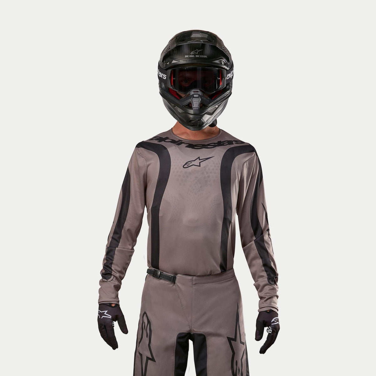 Motocross rider wearing the Alpinestars 2024 Fluid Lurv Jersey in Mud Black, an earthy grey-brown fabric with black graphic stripes and a star logo on the chest, paired with a black helmet, red goggles, and black gloves for a coordinated off-road racing look