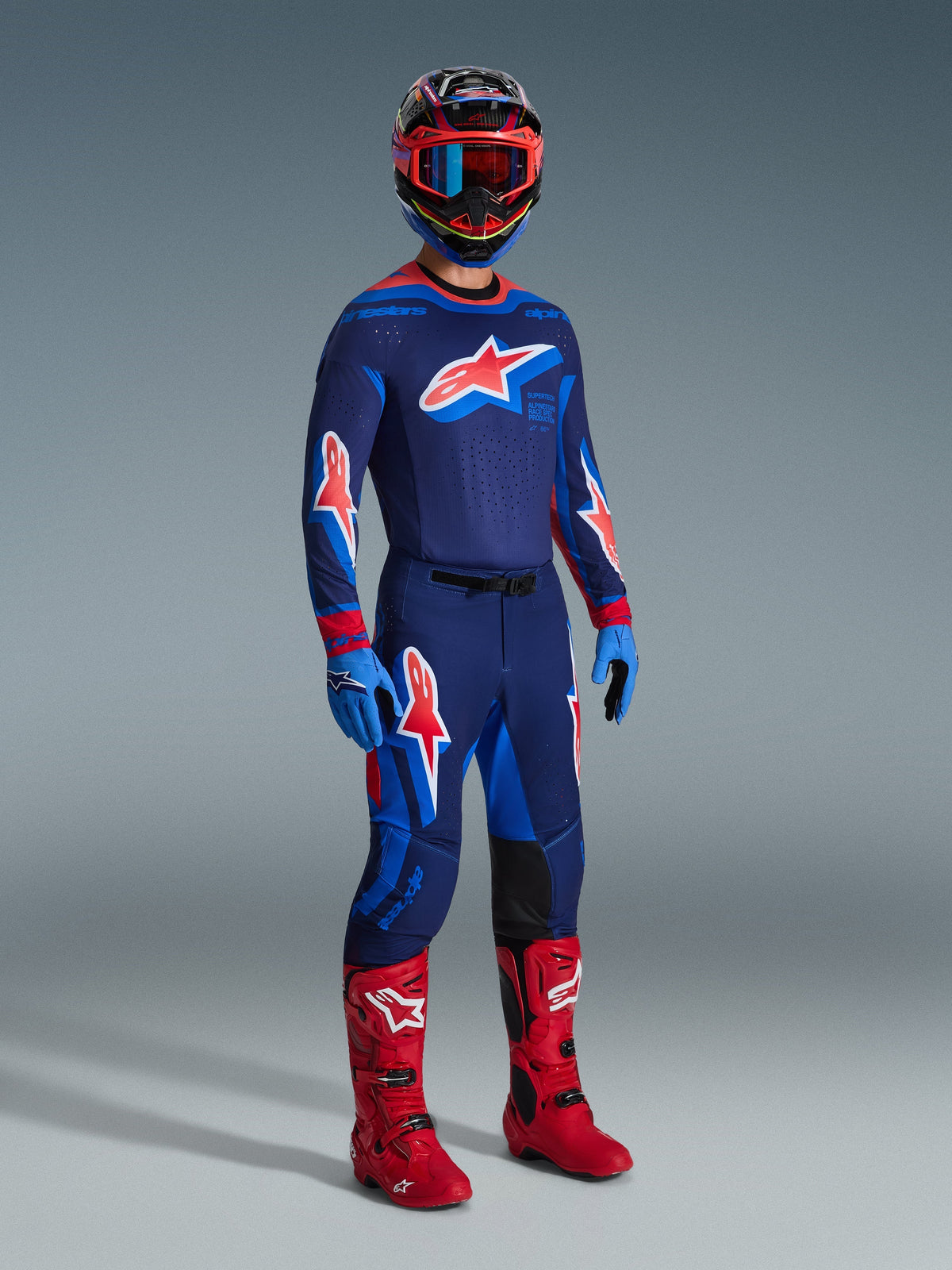 Motocross rider wearing complete racing gear, 2026 Supertech Vista Jersey in Dark Navy with Hot Coral and Cobalt accents, matching motocross pants, multi-colored helmet with red goggles, bright red boots, blue gloves, coordinated outfit for off-road racing