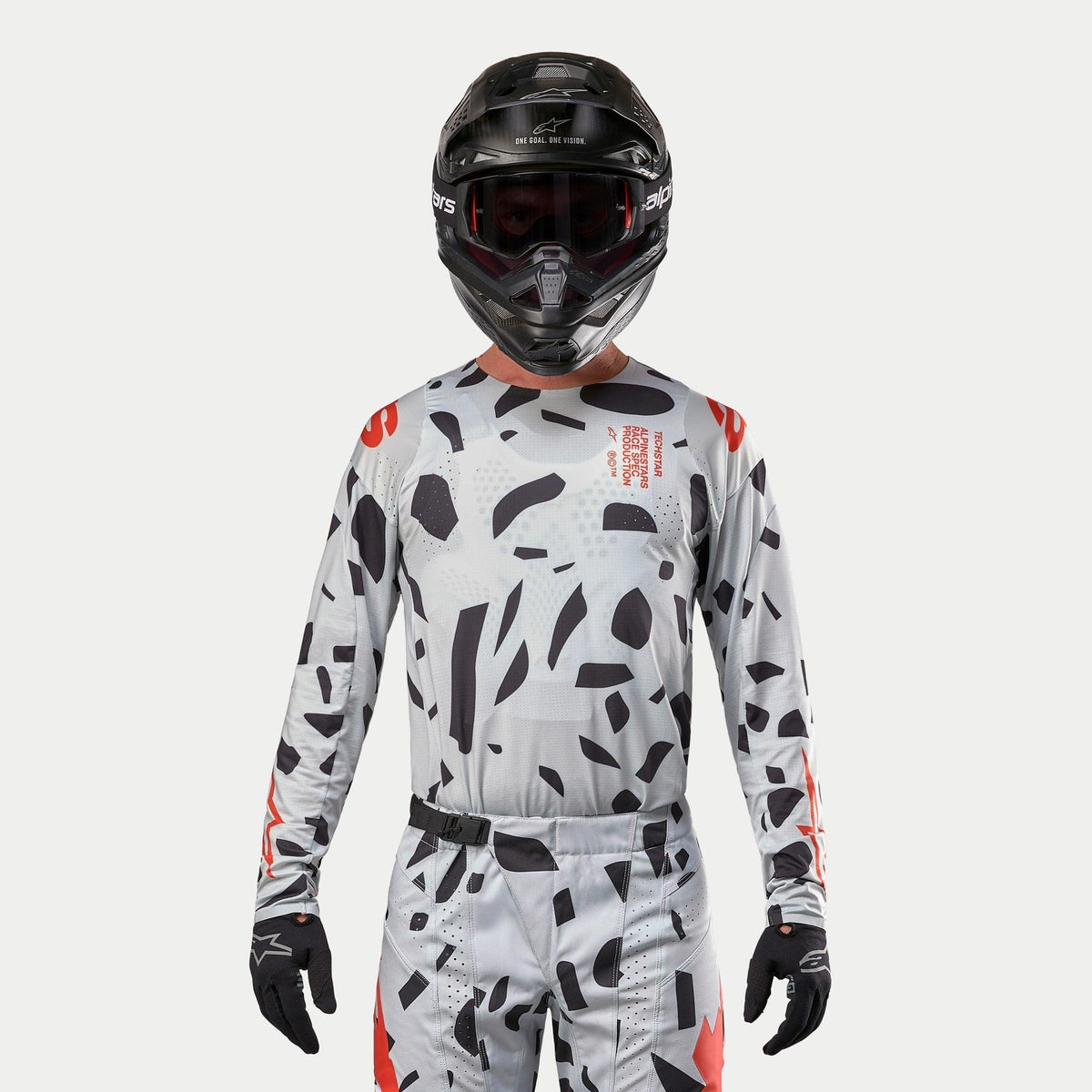 Alpinestars 2024 Techstar Rantera Trikot, Motocross-Trikot, Haze Gray Camo mit schwarzen unregelmäßigen Mustern und roten Akzenten, getragen von einem Fahrer mit passender Hose, schwarzem Helm und Handschuhen, Offroad-Rennausrüstung.