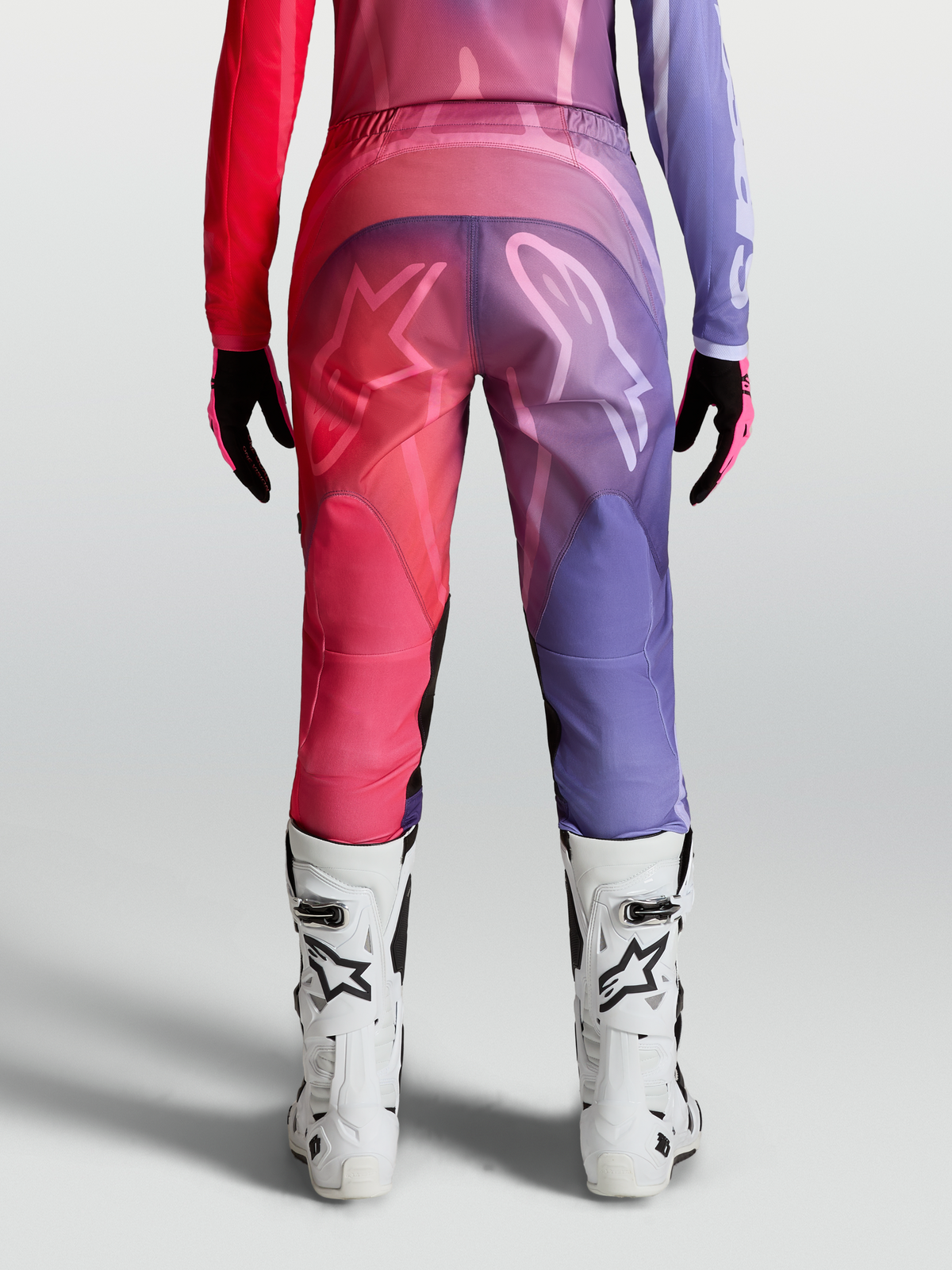 Alpinestars Stella Fluid Kepler Hose, Damen-Motocross-Hose in Lila und Rot, Rückansicht mit ergonomischem Schnitt, 600D-Polygewebe und weißen Logo-Grafiken, getragen mit weißen MX-Stiefeln.