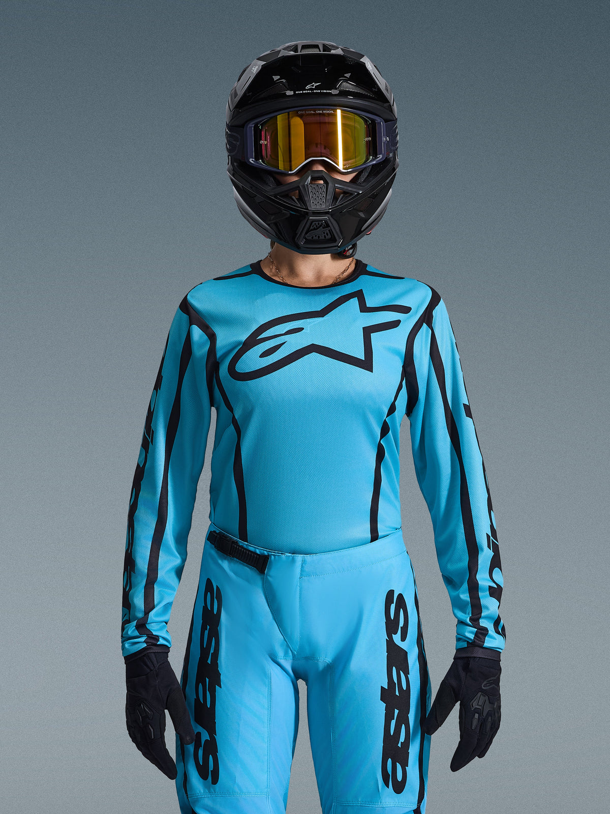 Motocross-Fahrerin im Alpinestars 2026 Stella Fluid Apex Bekleidungsset für Damen, hellblaues Trikot mit schwarzen Konturen und Logo, passende Hose, schwarzer Helm mit irisierender goldener Brille und schwarze Handschuhe, abgestimmtes Motocross-Rennyoutfit für Damen