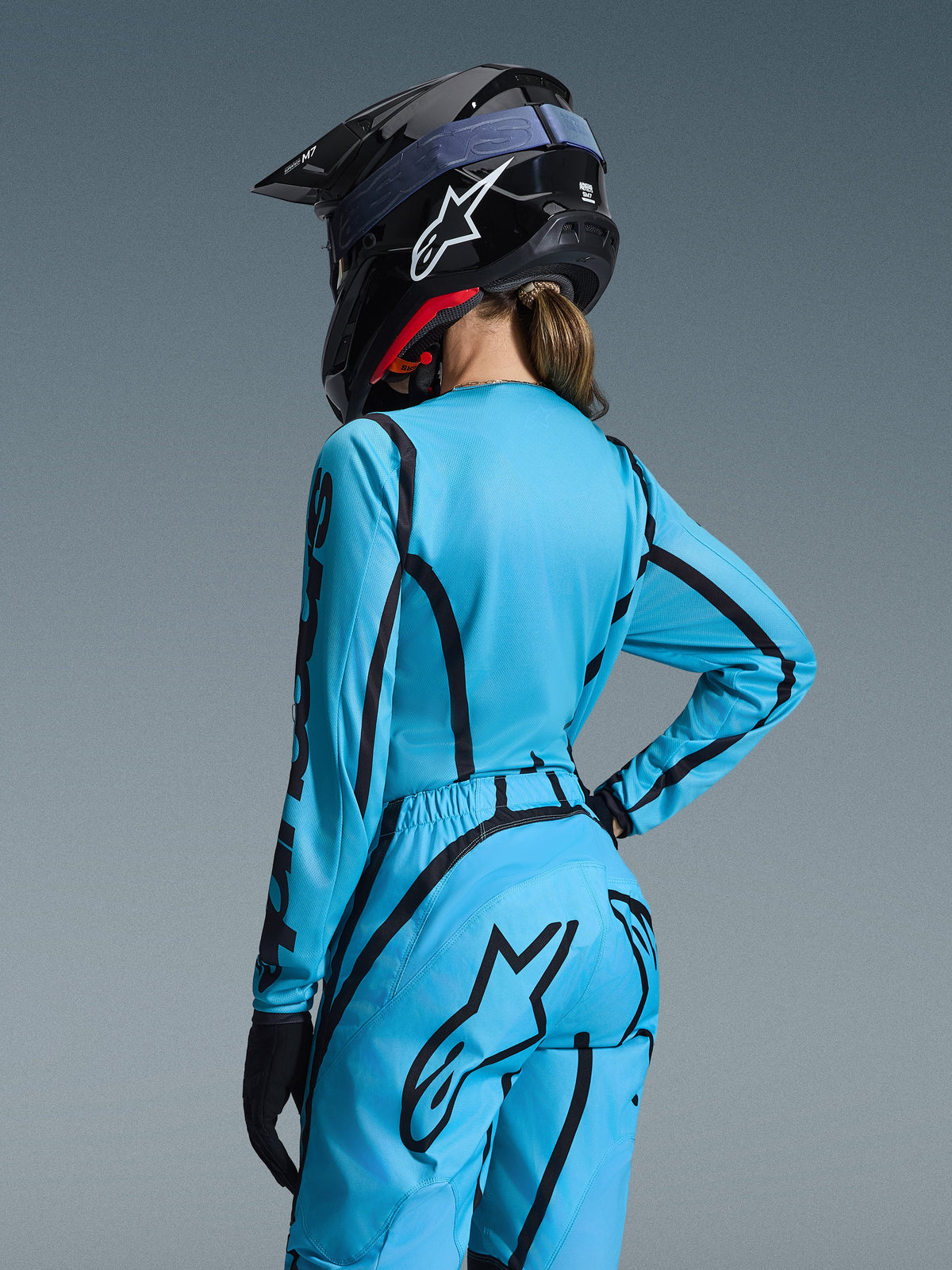 Alpinestars 2026 Stella Fluid Apex Gear Set, Hellblau und Schwarz, Rückansicht einer Fahrerin in Motocross-Trikot und passender Hose, schwarzer Alpinestars-Helm mit weißem Logo, technische Offroad-Rennbekleidung für Damen.
