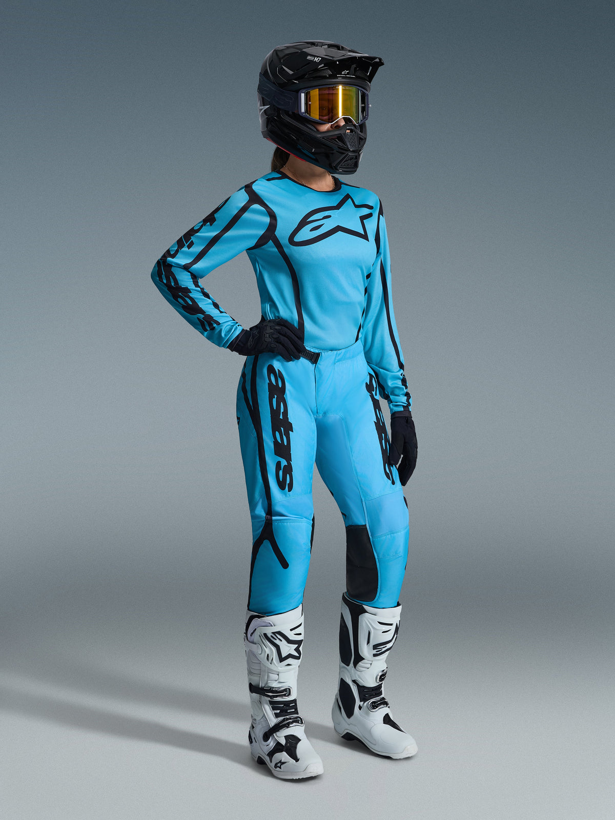 Alpinestars 2026 Stella Fluid Apex Ausrüstungsset für Damen in Tropical Black, bestehend aus einem hellblauen Trikot und einer passenden Hose mit schwarzen Akzenten, getragen von einer Fahrerin mit schwarzem Helm, irisierender Brille und weißen Stiefeln, professionelle Motocross-Rennausrüstung.