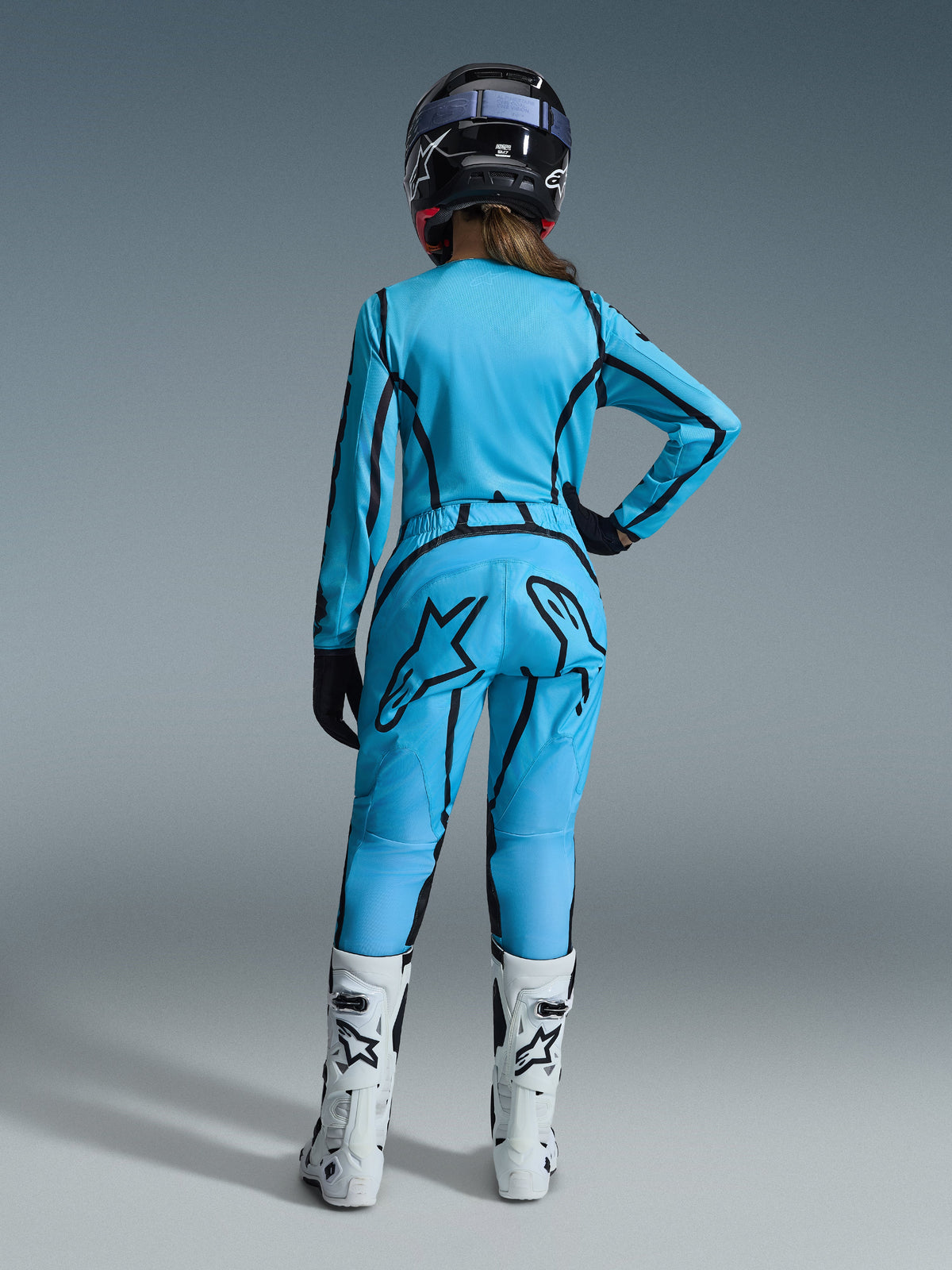 Motocross-Fahrerin in der Rückansicht, trägt das Alpinestars 2026 Woman Stella Fluid Apex Gear Set in leuchtendem Blau mit schwarzen grafischen Akzenten, ein großes schwarzes Stern-Logo auf der Hose, kombiniert mit weiß-schwarzen Stiefeln und einem schwarzen Helm.