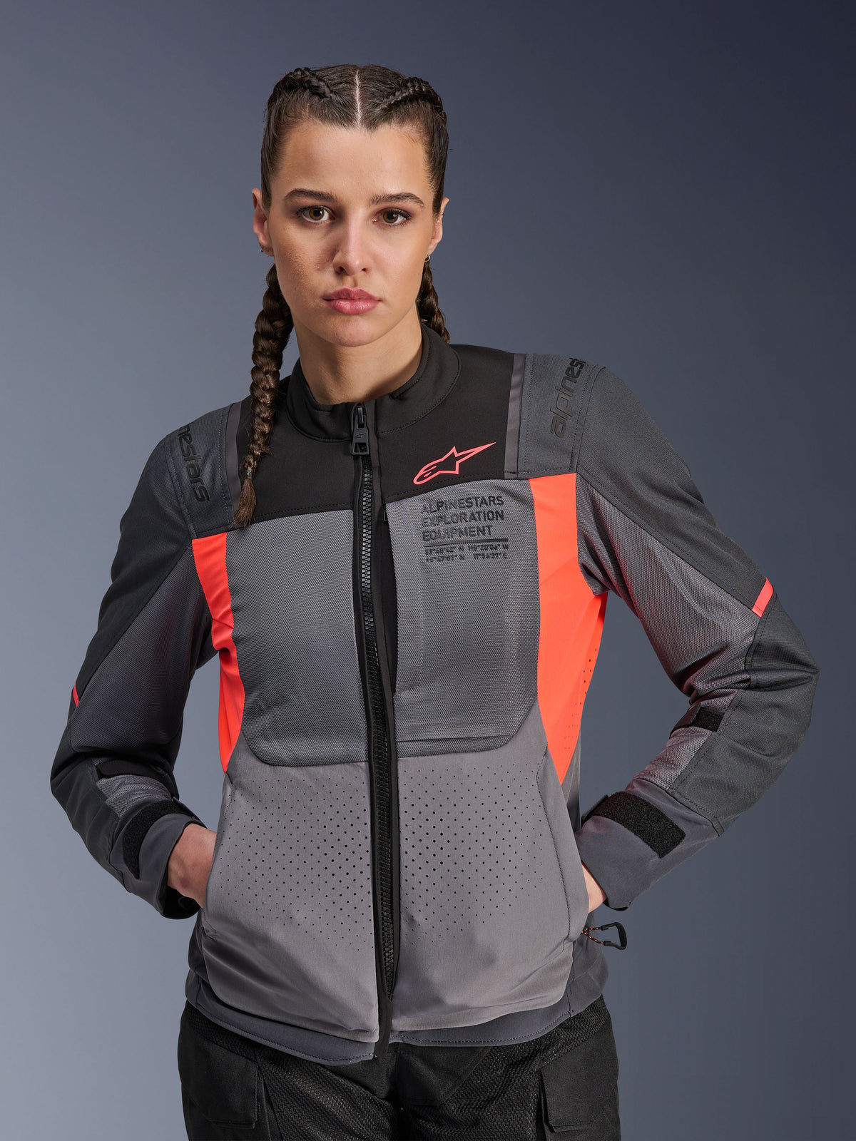 Alpinestars Stella ST-2 Air Jacke, Damen-Motorradjacke, Deep Gray und Fluo Coral, getragen von Model mit geflochtenem Haar, mit Tech-Mesh-Einsätzen für Luftzirkulation, Laserperforationen und „Alpinestars Exploration Equipment“-Branding auf der Brust, Sommer-Tourenausrüstung