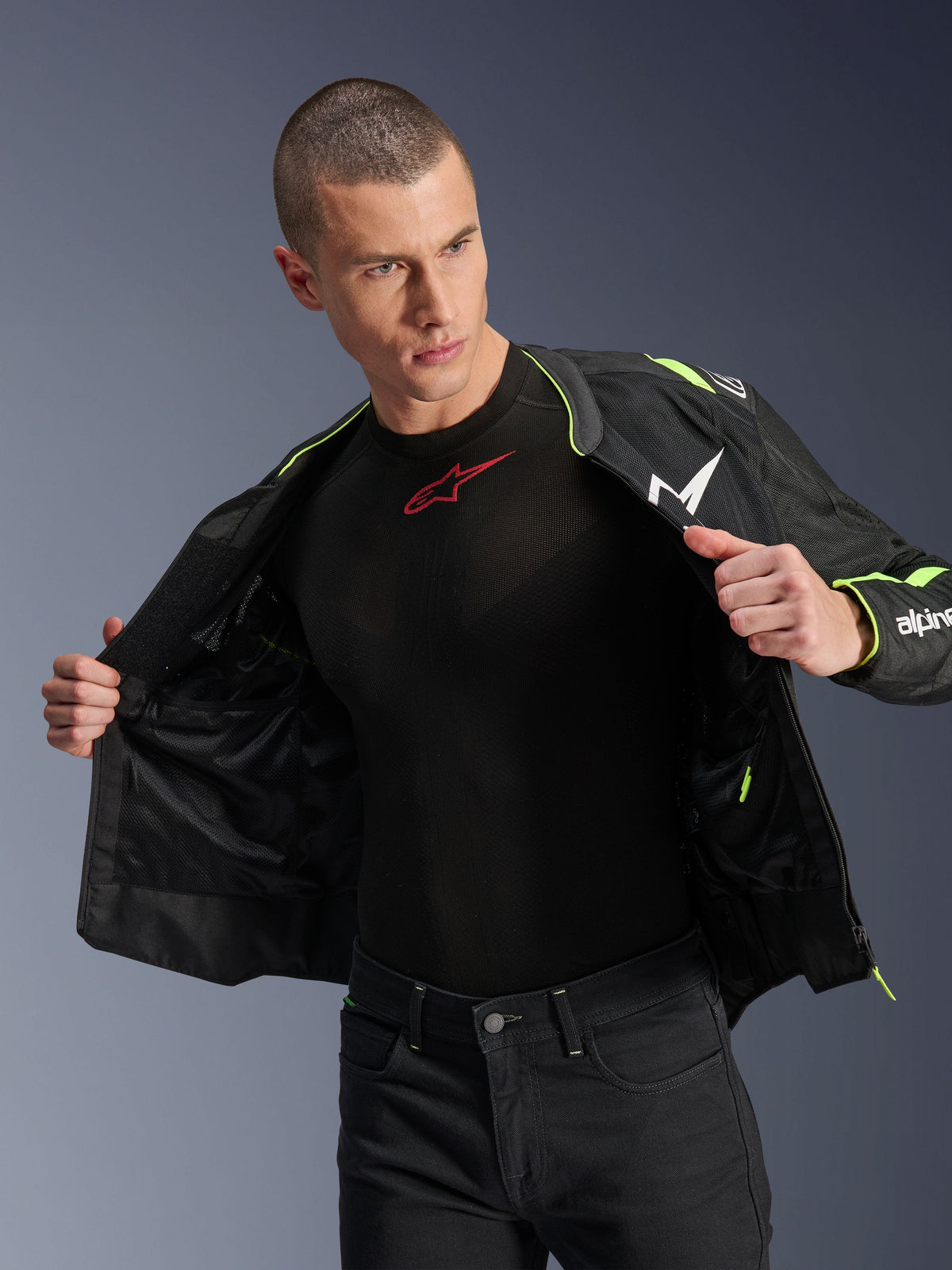 Männliches Model trägt die Alpinestars T-Sps Air V2 Jacke in Schwarz und Yellow Fluo, offen gezeigt, um die interne Mesh-Konstruktion und Taschen hervorzuheben, kombiniert mit einem schwarzen Alpinestars Base Layer und einer schwarzen Hose, urbaner Motorrad-Riding-Stil