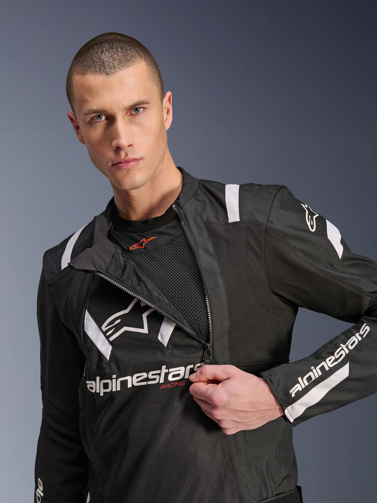 Alpinestars T-Stunt Air Jacke, Motorradjacke in Schwarz und Weiß, männliches Model zeigt teilweise geöffnetes modulares Frontpanel mit sichtbaren Mesh-Lagen zur Belüftung, weiße Alpinestars-Logos auf Brust und Ärmeln, sportliches Design für den Straßeneinsatz