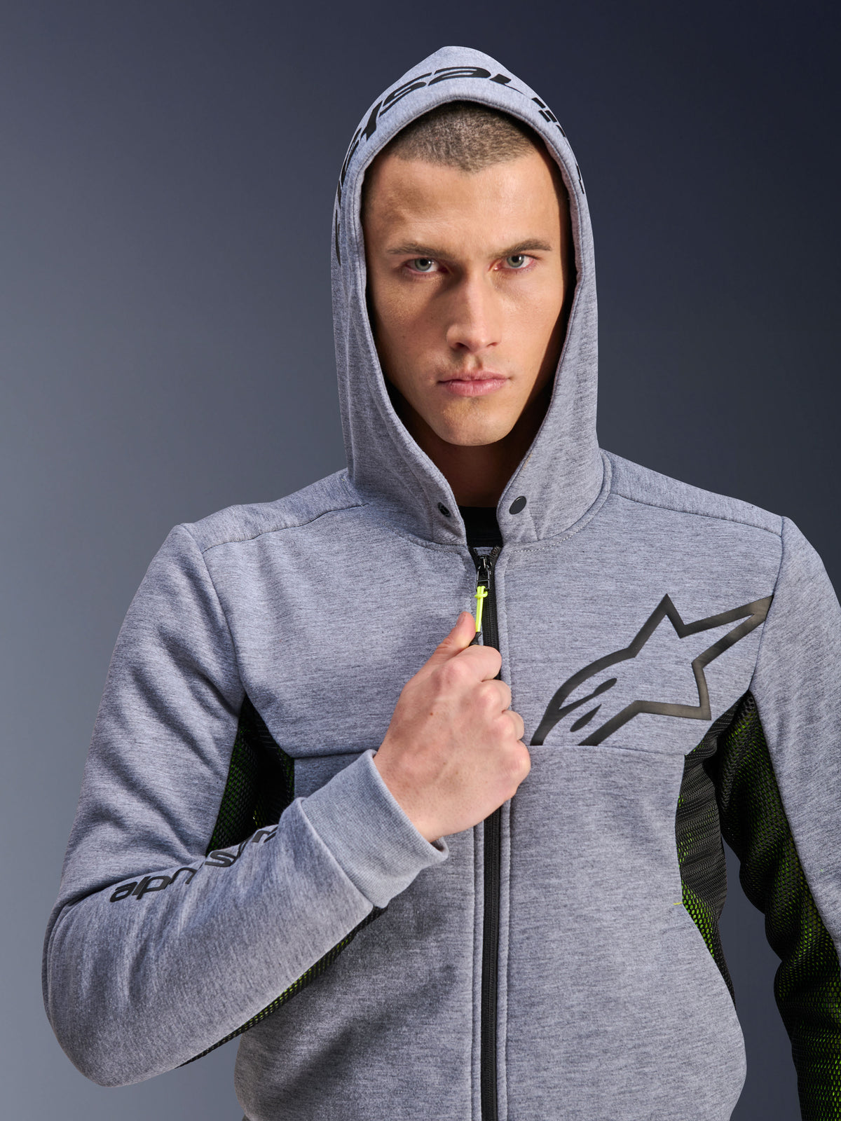 Alpinestars Chrome Air Sport Hoodie, Grau Meliert Schwarz, Motorrad-Hoodie getragen von einem Mann mit aufgesetzter Kapuze, mit schwarzem Stern-Logo auf der Brust, neongelbem Reißverschluss-Zipper und seitlichen Mesh-Einsätzen zur Belüftung, urbaner Fahrstil