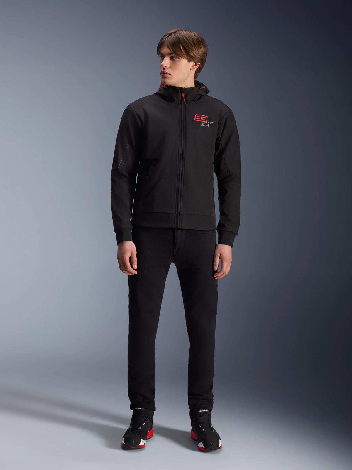 Alpinestars MM93 Radium Tech Hoodie, schwarzer urbaner Motorrad-Hoodie, mit rotem 93-Logo und weißem Stern-Logo auf der Brust, getragen von einem Model mit schwarzer Hose und passenden Motorradschuhen, elegante winddichte technische Shell für das urbane Fahren.