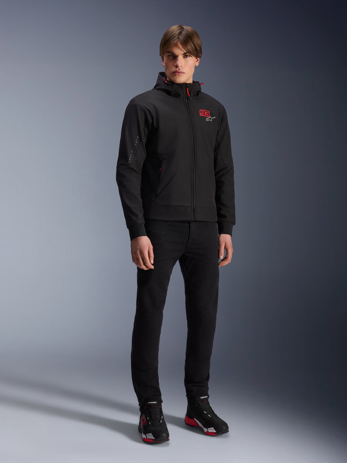 Alpinestars MM93 Radium Tech Hoodie, urbane Motorradjacke, Schwarz mit leuchtend roten Akzenten, getragen von einem Model mit schwarzer Hose und Motorradschuhen, mit rotem „93“-Logo auf der Brust und Marc Marquez Branding am Ärmel, lässige technische Streetwear für die Stadt