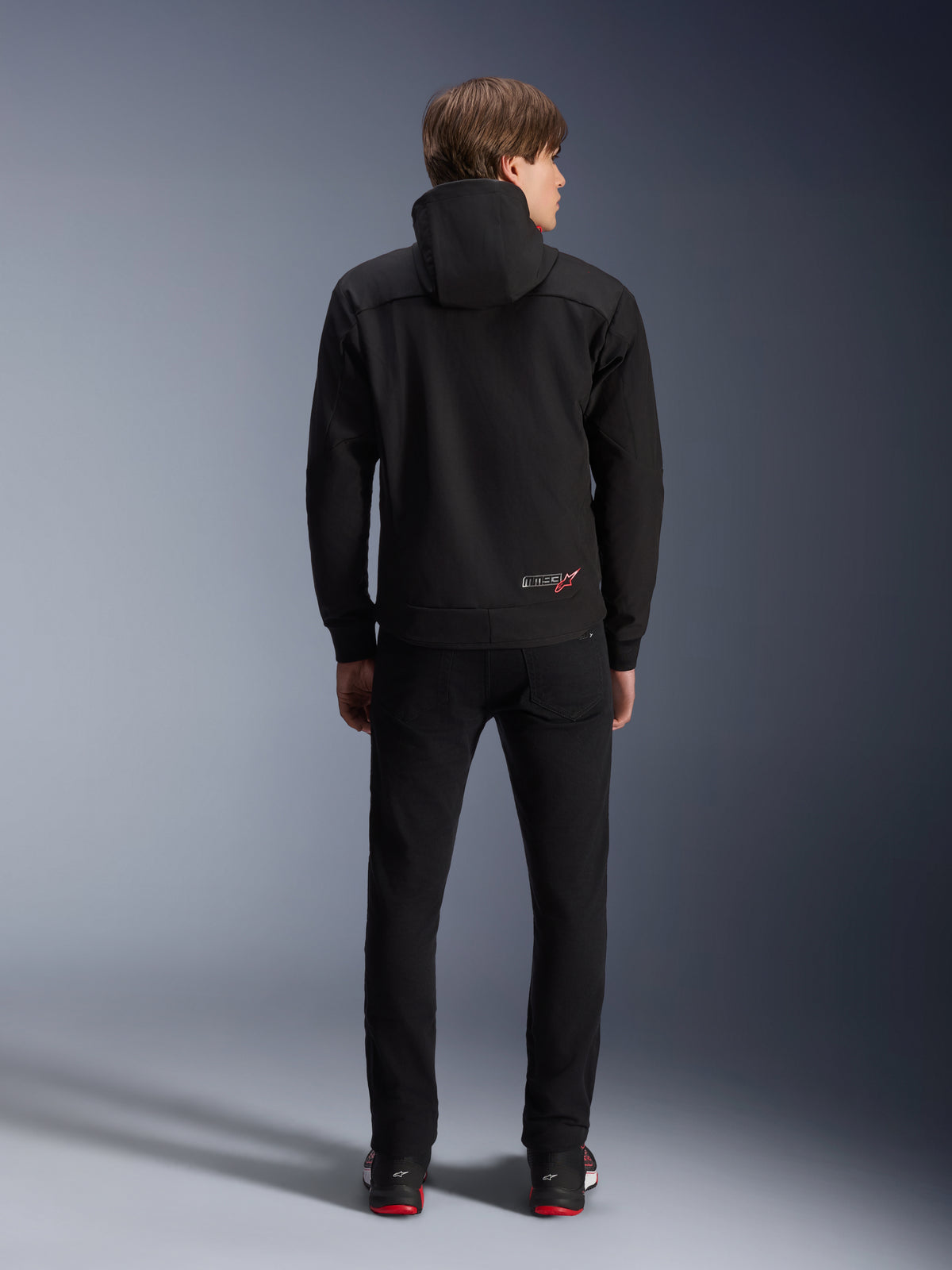 Alpinestars MM93 Radium Tech Hoodie, Schwarz, Rückansicht am Model, mit kleinem MM93-Logo mit rotem Stern am unteren Rücken, kombiniert mit schwarzer Hose, urbane Motorradbekleidung
