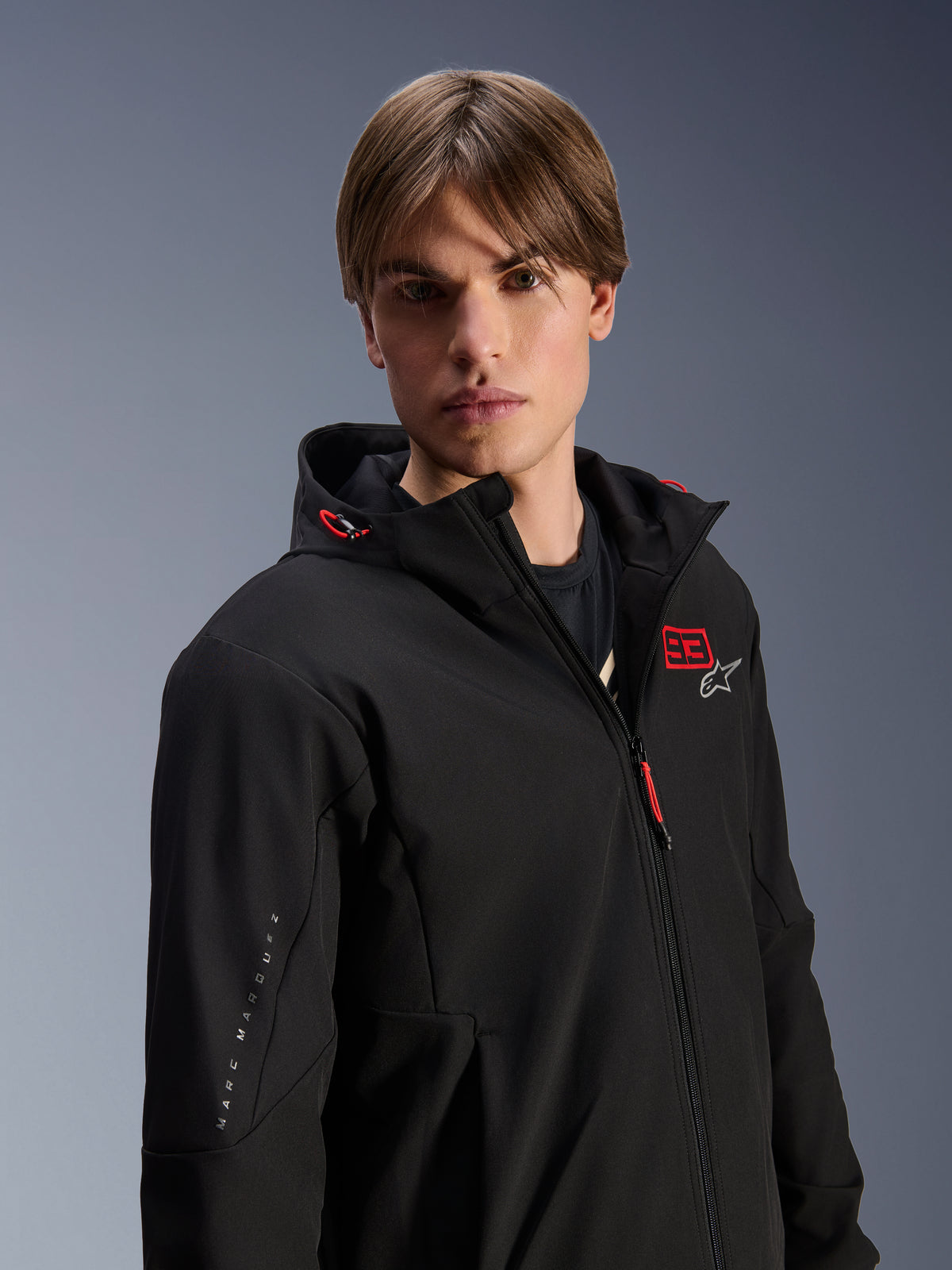 Männliches Modell im Alpinestars MM93 Radium Tech Hoodie, schwarze urbane Motorradjacke, rotes '93'-Logo und weißes Stern-Logo auf der Brust, 'Marc Marquez'-Branding am Ärmel, technisches winddichtes Material für urbanes Fahren.